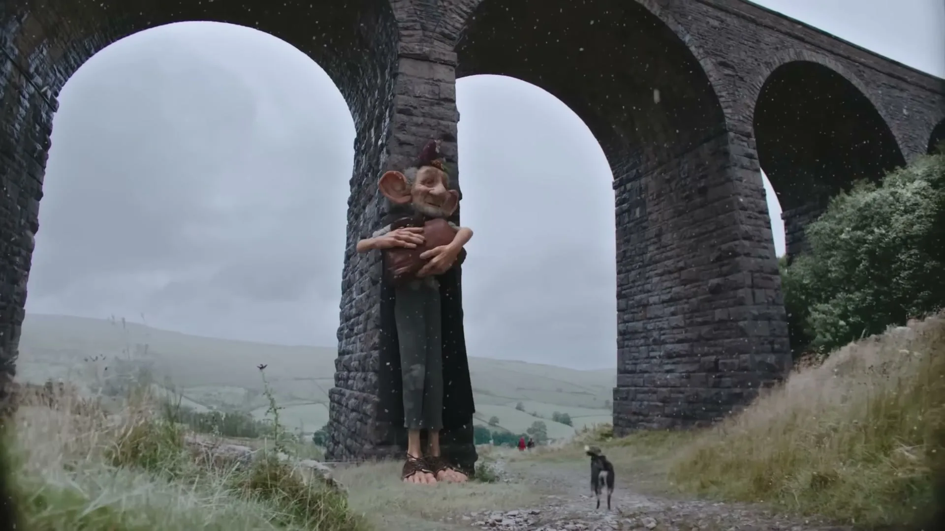 A giant, troll-like creature with large ears and a small bird on its head stands under a stone archway, holding a brown object, as a small dog walks nearby in a rural landscape.