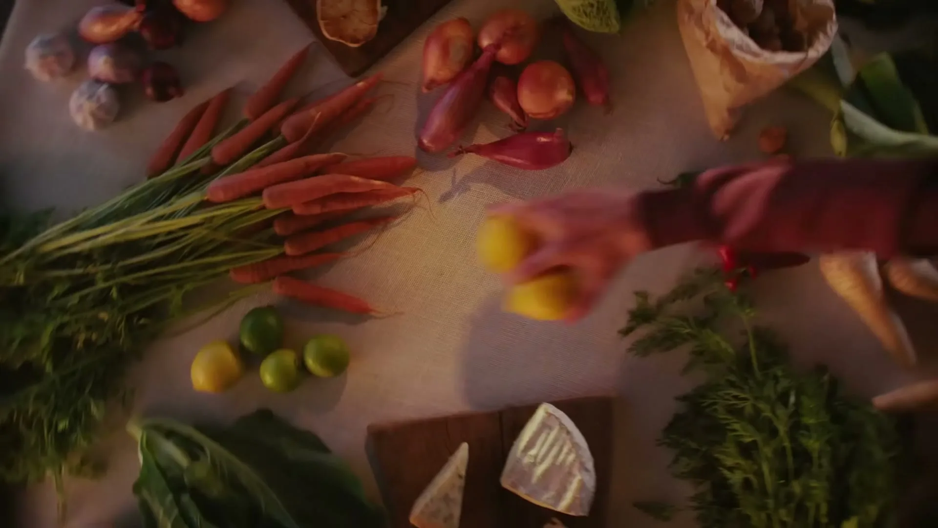 A person's hands arrange a variety of fresh vegetables, cheeses, and herbs on a rustic wooden table, preparing for a meal.