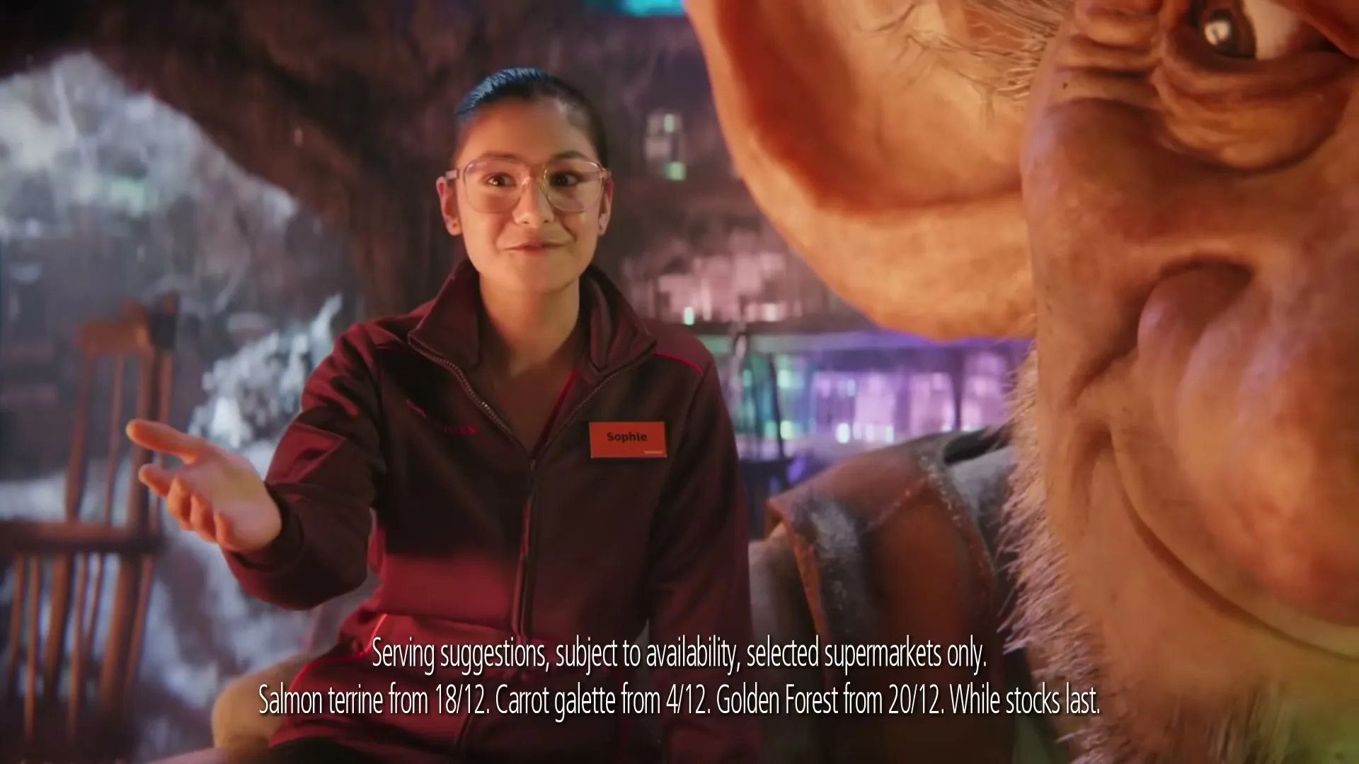 A young female Sainsbury's employee in glasses and a red jacket talks to a large, friendly-looking CGI creature in a fantastical, dimly lit cave-like setting.