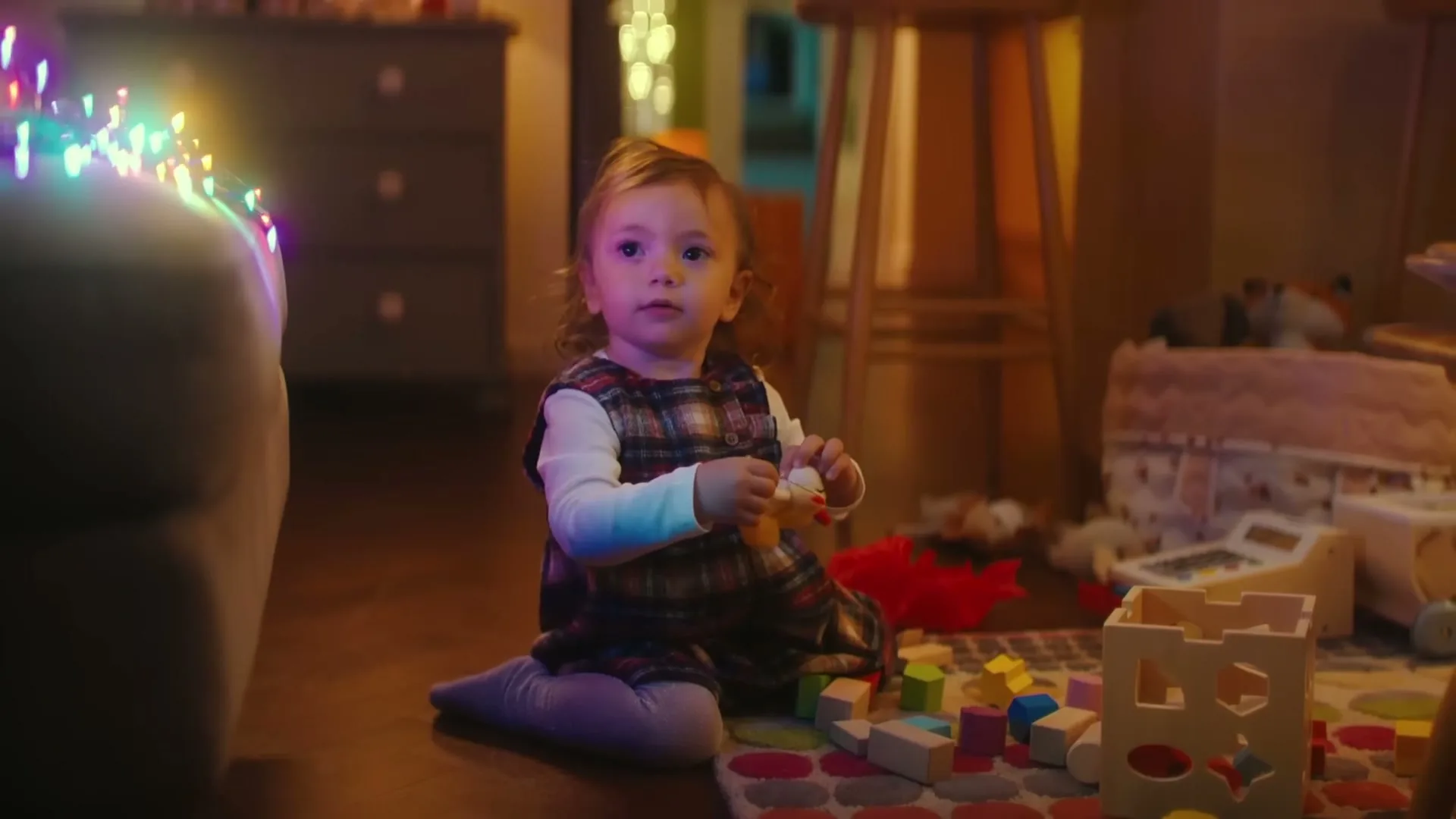 A cute baby girl with light brown hair, wearing a plaid dress, sits on the floor playing with colorful blocks and a toy in a cozy living room decorated with Christmas lights.