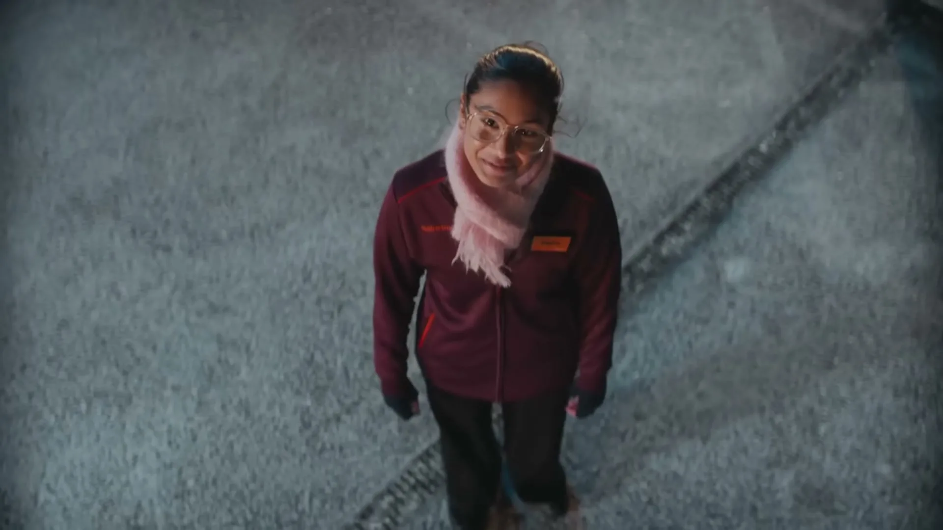 A young female Sainsbury's employee in a maroon jacket and pink scarf smiles up at the camera as snow falls around her at night.
