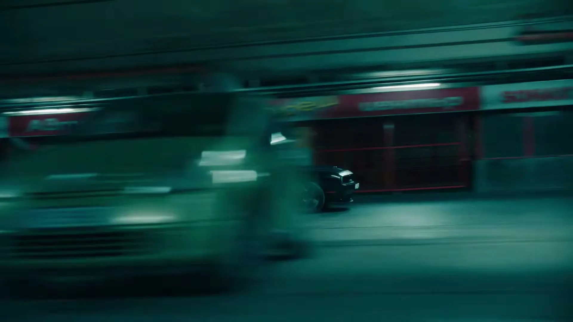 A black car with a red stripe drives quickly through a dimly lit underground parking garage, with other vehicles blurred in the background.
