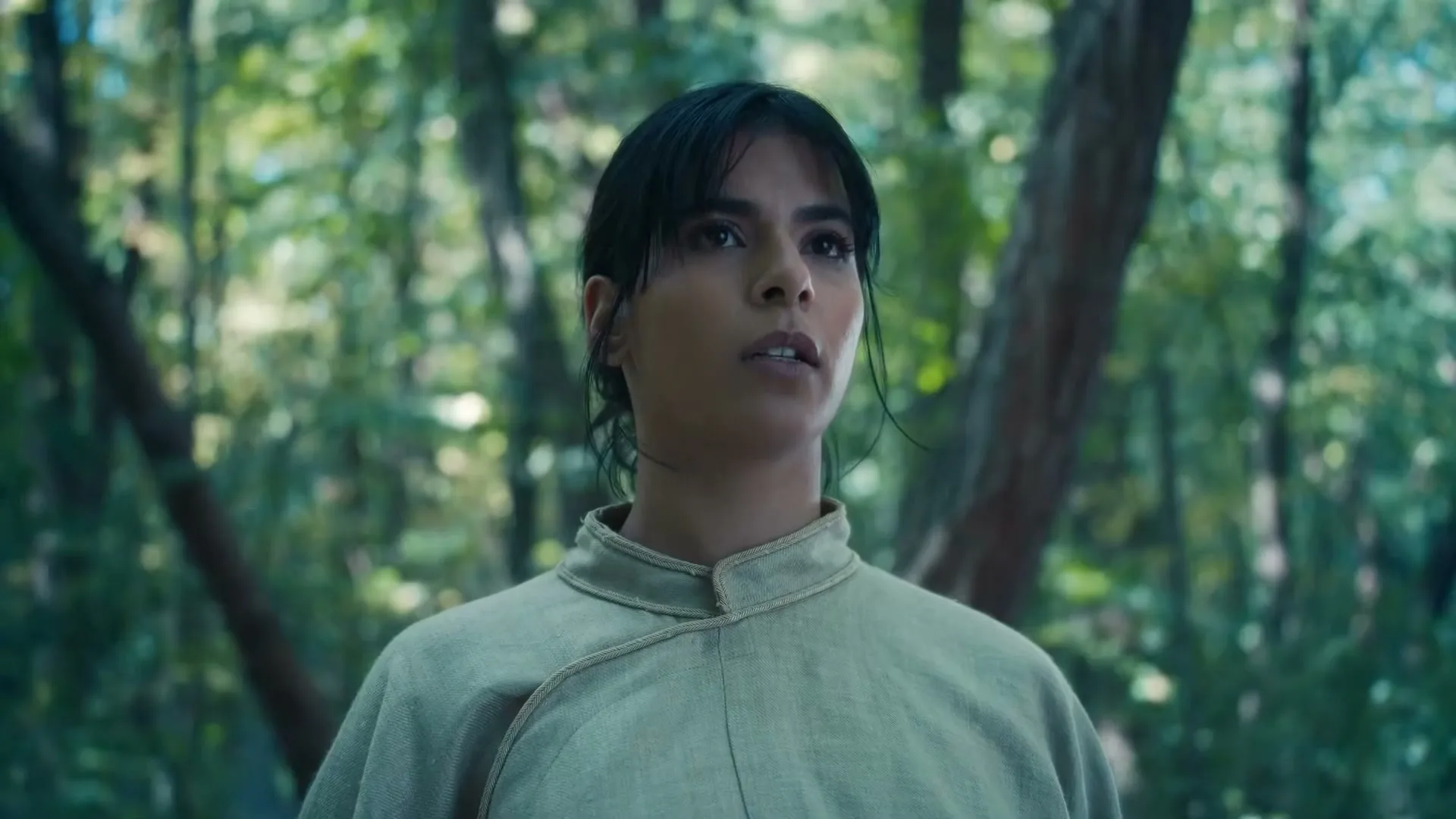 A young woman with dark hair and a light-colored tunic stands in a lush green forest, looking up with a concerned expression.