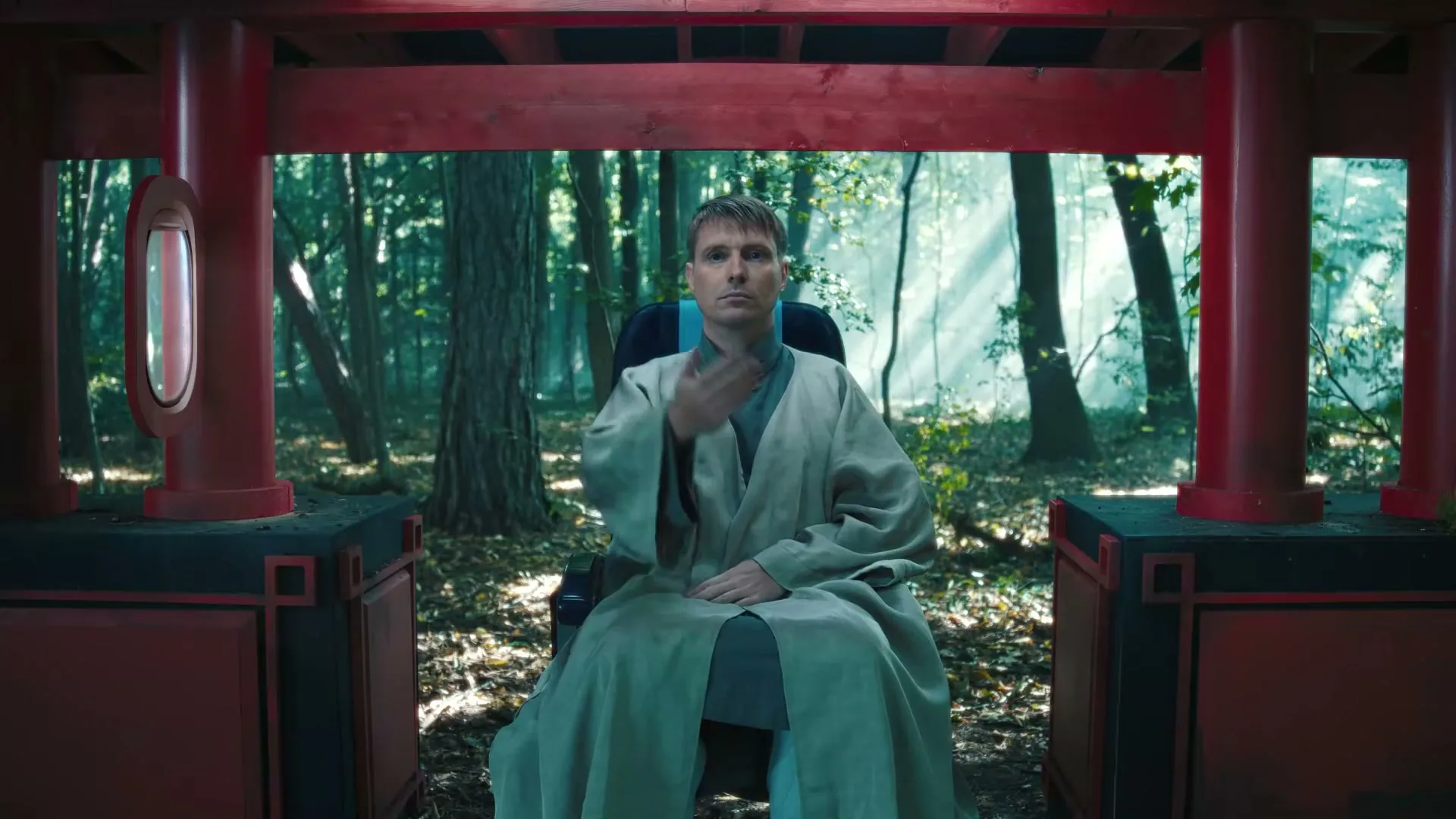 A man in a robe sits in a chair, framed by a red structure, in a sun-dappled forest, gesturing with his hand.