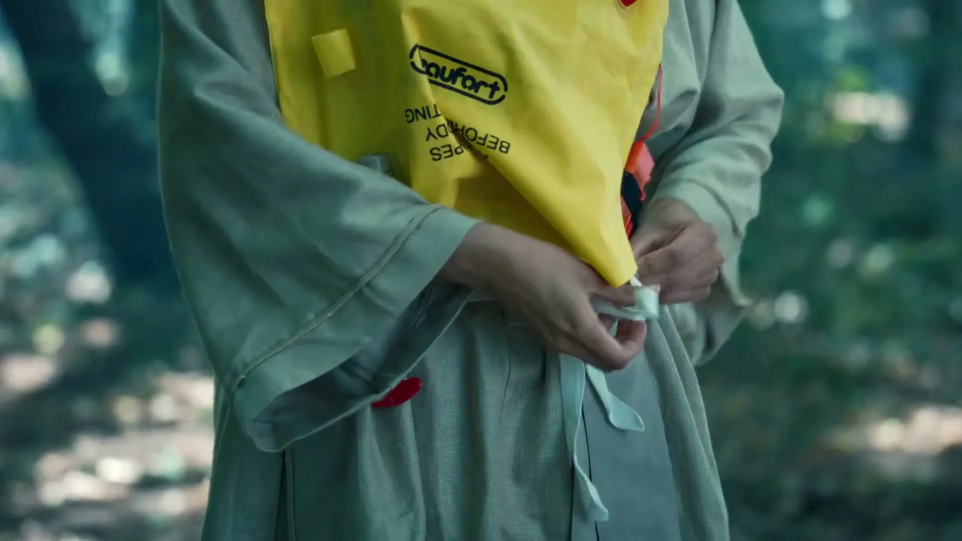 A person in a beige robe ties a yellow life vest around their waist in a forest setting.