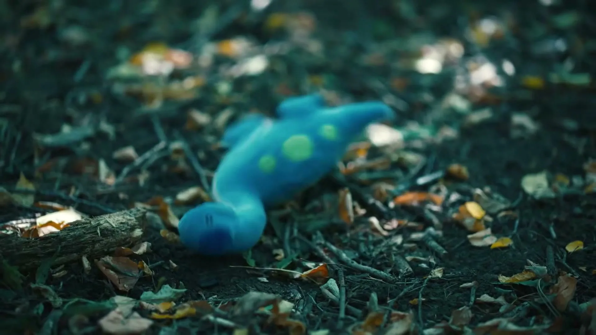 A blue stuffed animal with green spots falls onto a forest floor covered in leaves and twigs.
