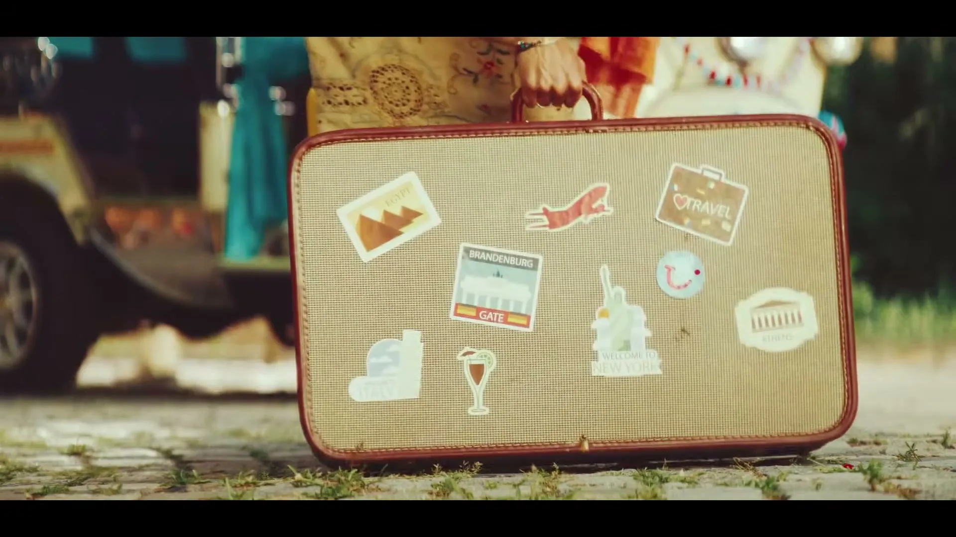 A person in traditional clothing and sandals holds a vintage suitcase covered in travel stickers, standing on a cobblestone path next to a colorful vehicle.