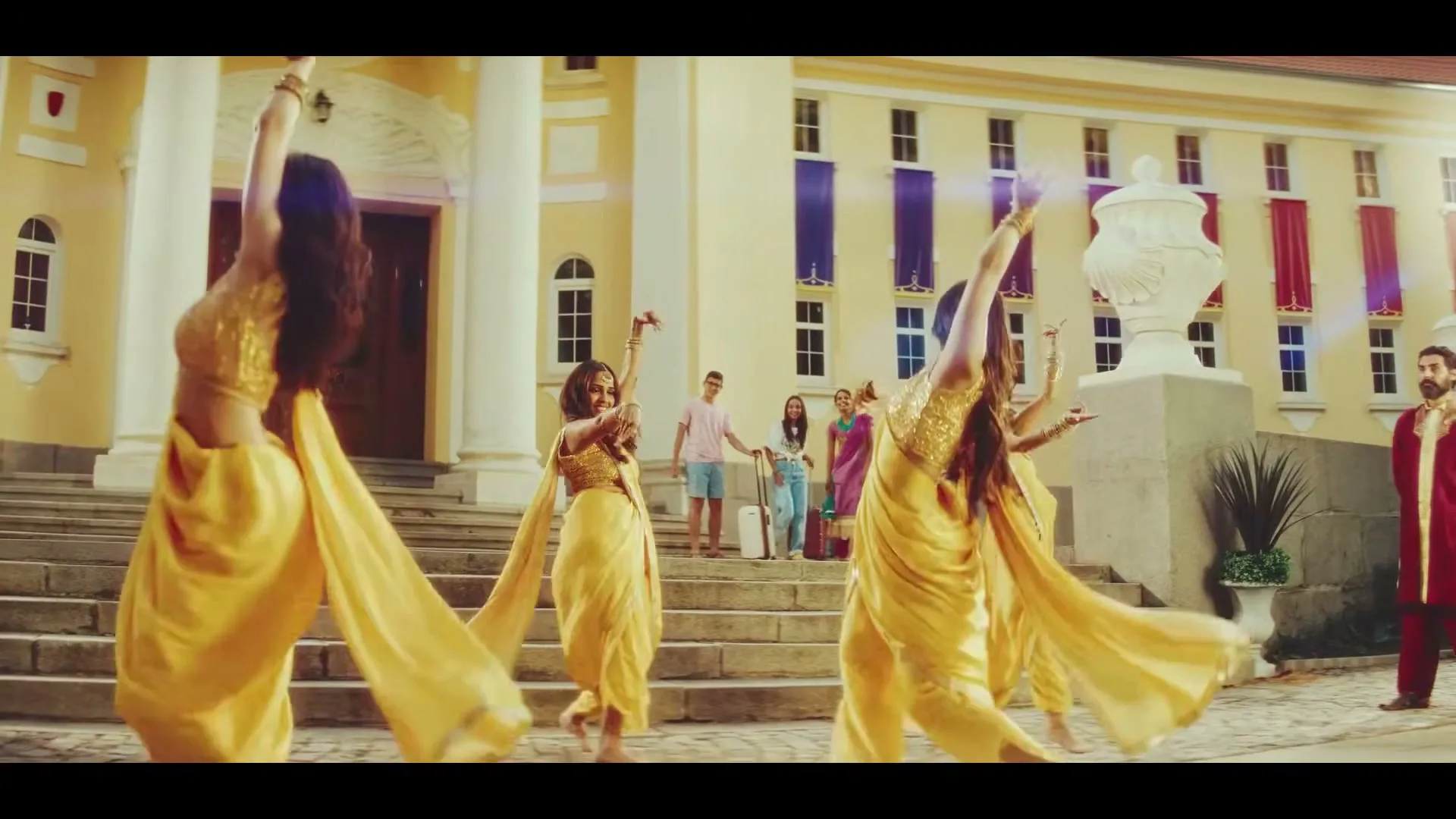 Three women in yellow saris dance in front of a grand building, while other people watch from the steps, creating a festive atmosphere.