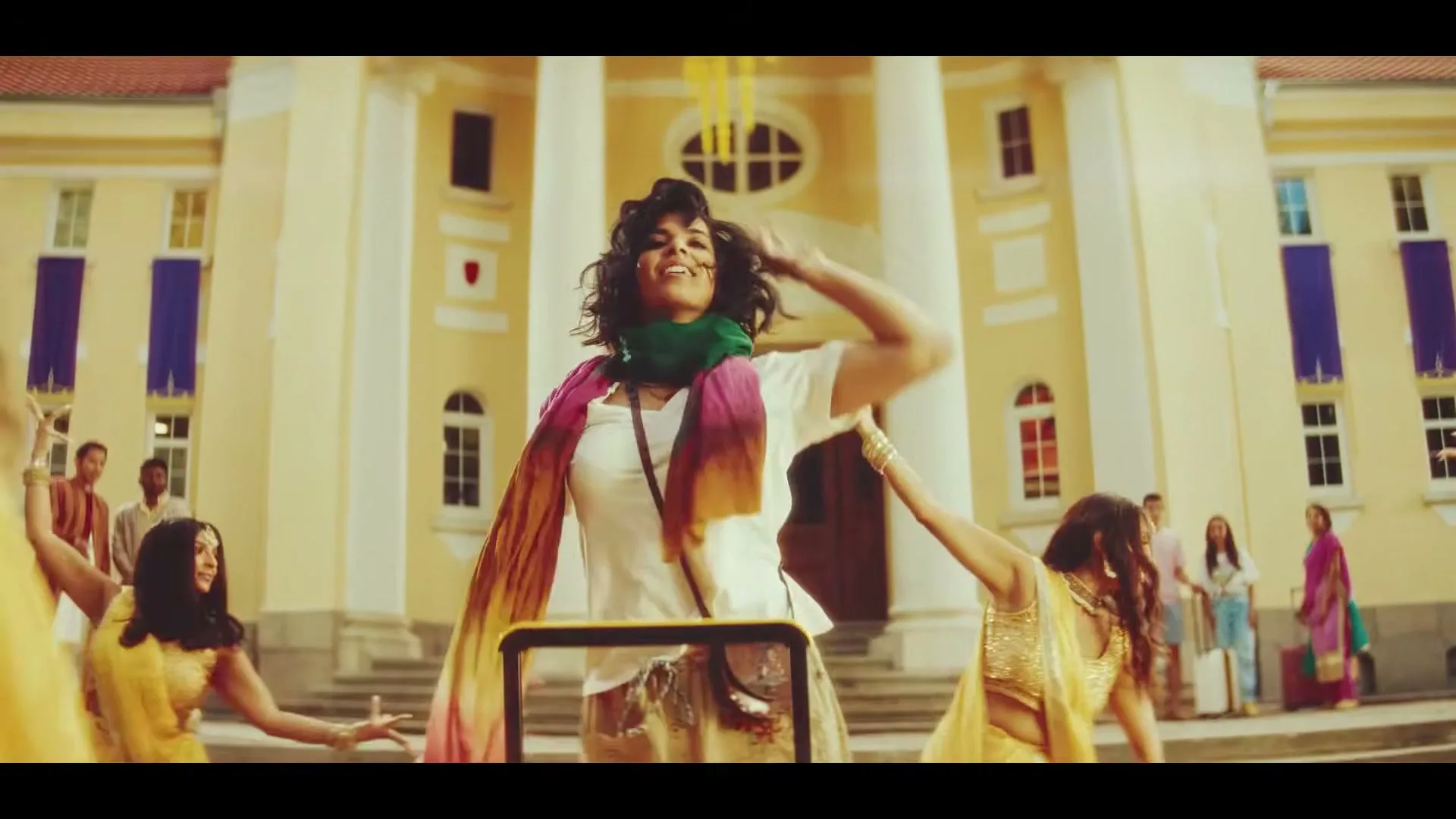 A diverse group of people, some in traditional Indian attire, dance joyfully in front of a grand, light-colored building with columns and steps.