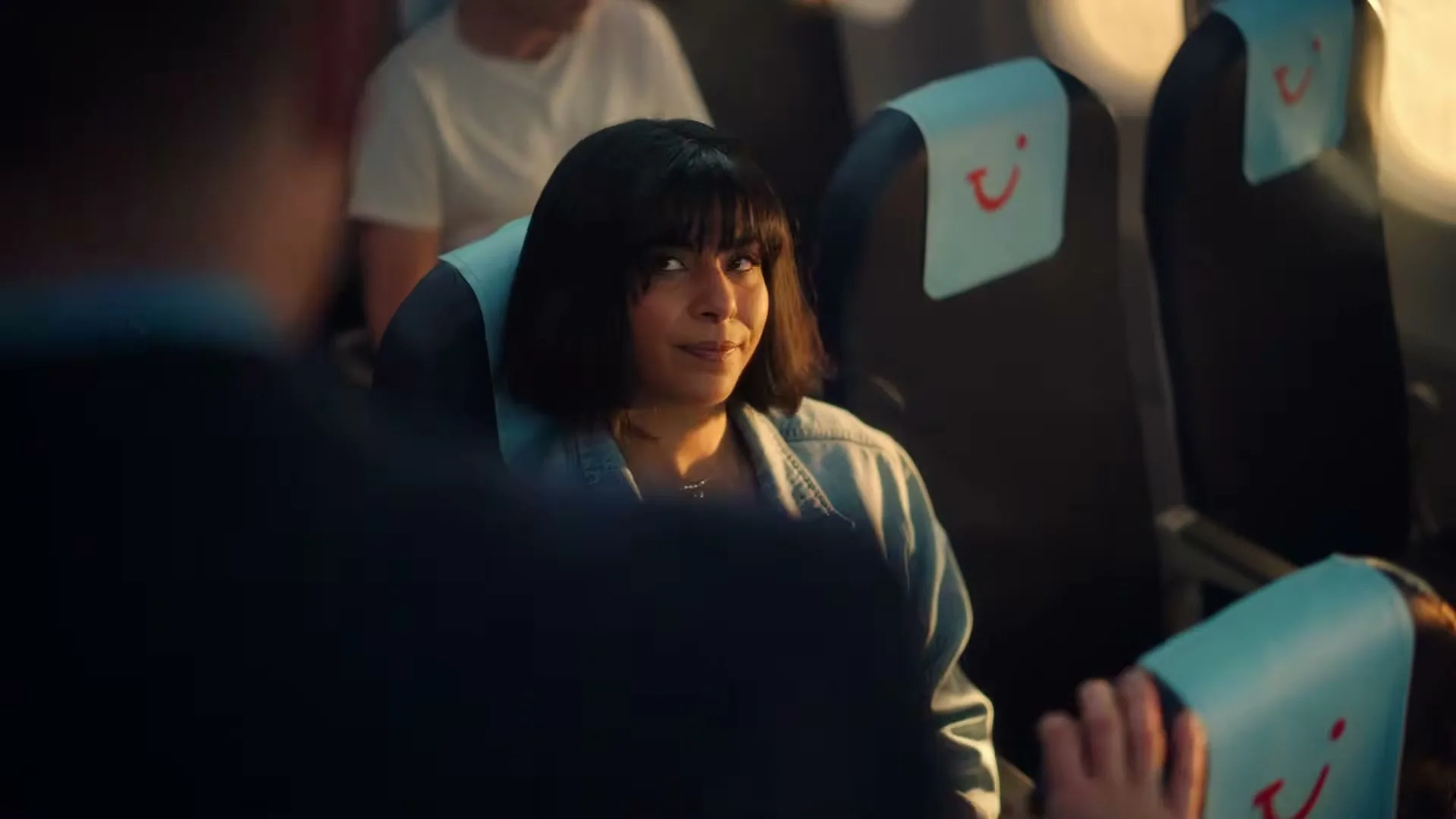 A young woman with dark hair and a denim jacket sits in an airplane seat, looking around with a curious expression as a man's arm is visible in the foreground.