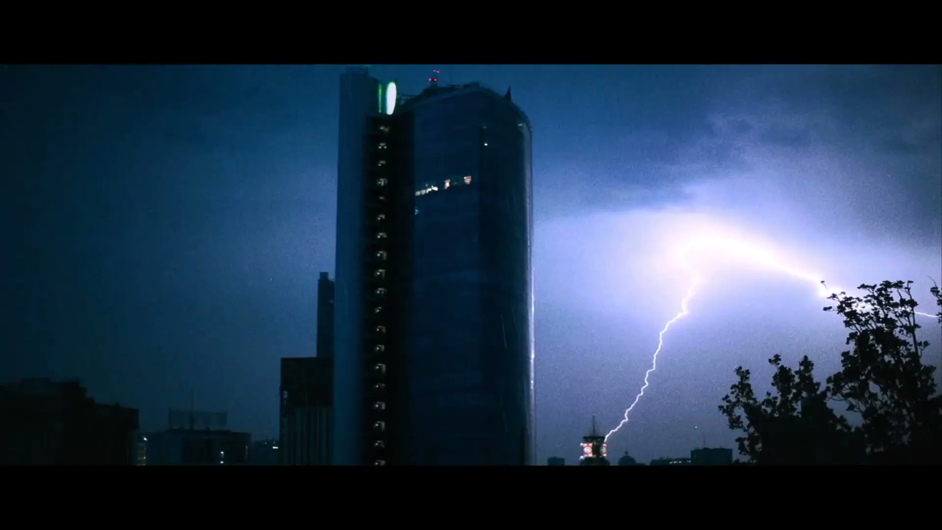 A tall, modern skyscraper stands against a dark, stormy night sky as lightning flashes across the frame.