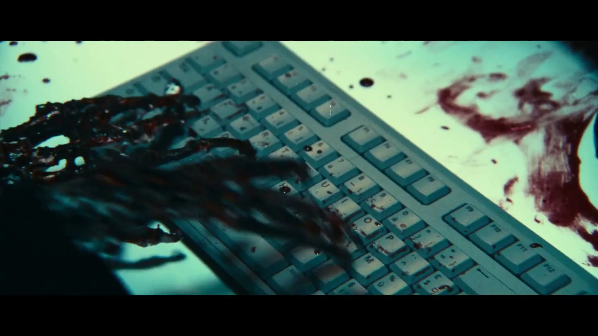 A bloody, skeletal hand types on a computer keyboard, with blood splattered across the desk in a dimly lit office.