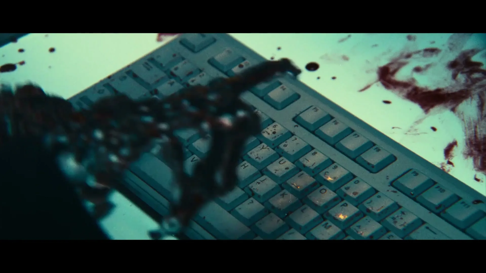 A skeletal hand, covered in blood, types on a dirty, blood-splattered computer keyboard in a dimly lit room.