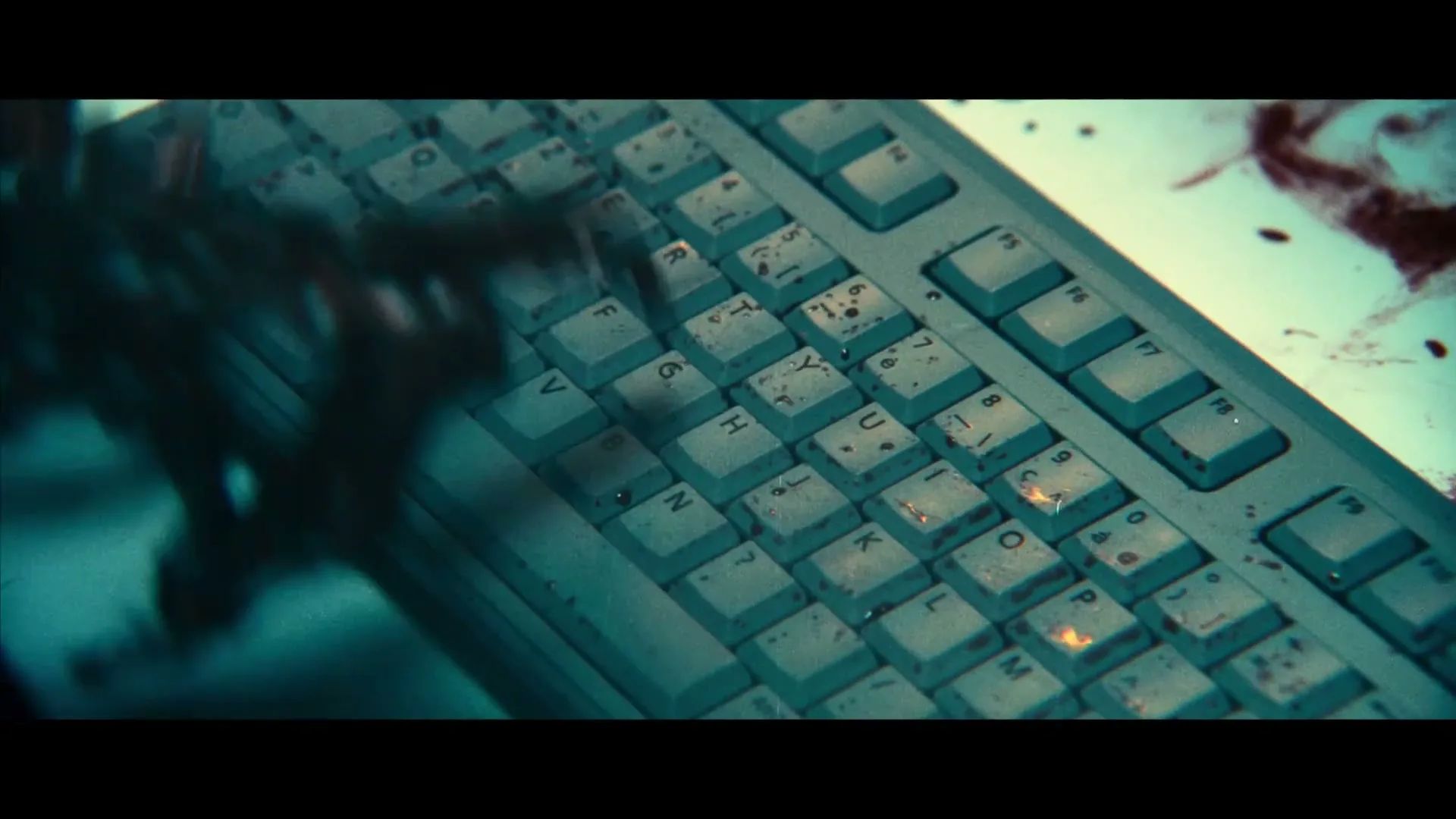 A dark, shadowy hand with long fingers types on a blood-splattered computer keyboard in a dimly lit room.