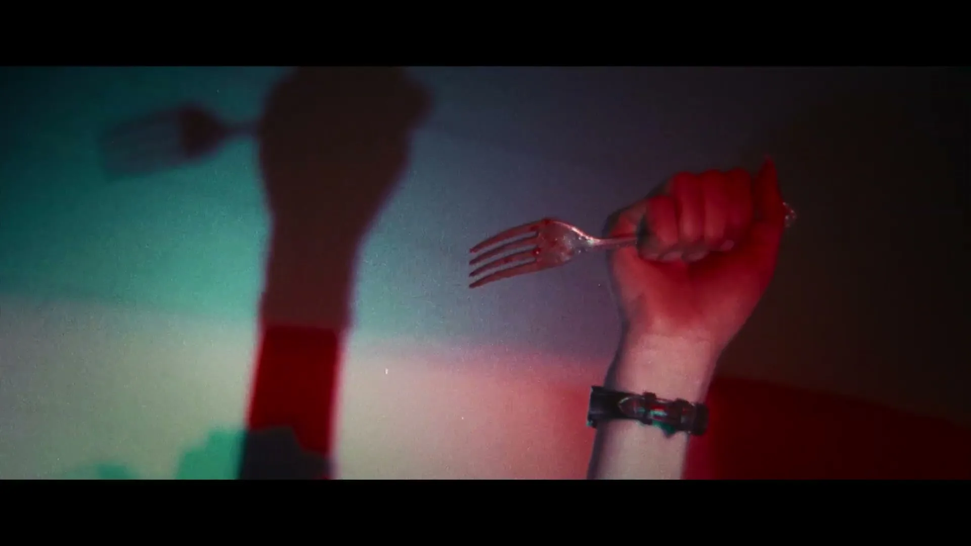 A person's arm with a spiked bracelet holds a fork, casting a long shadow on a wall with colorful lighting.