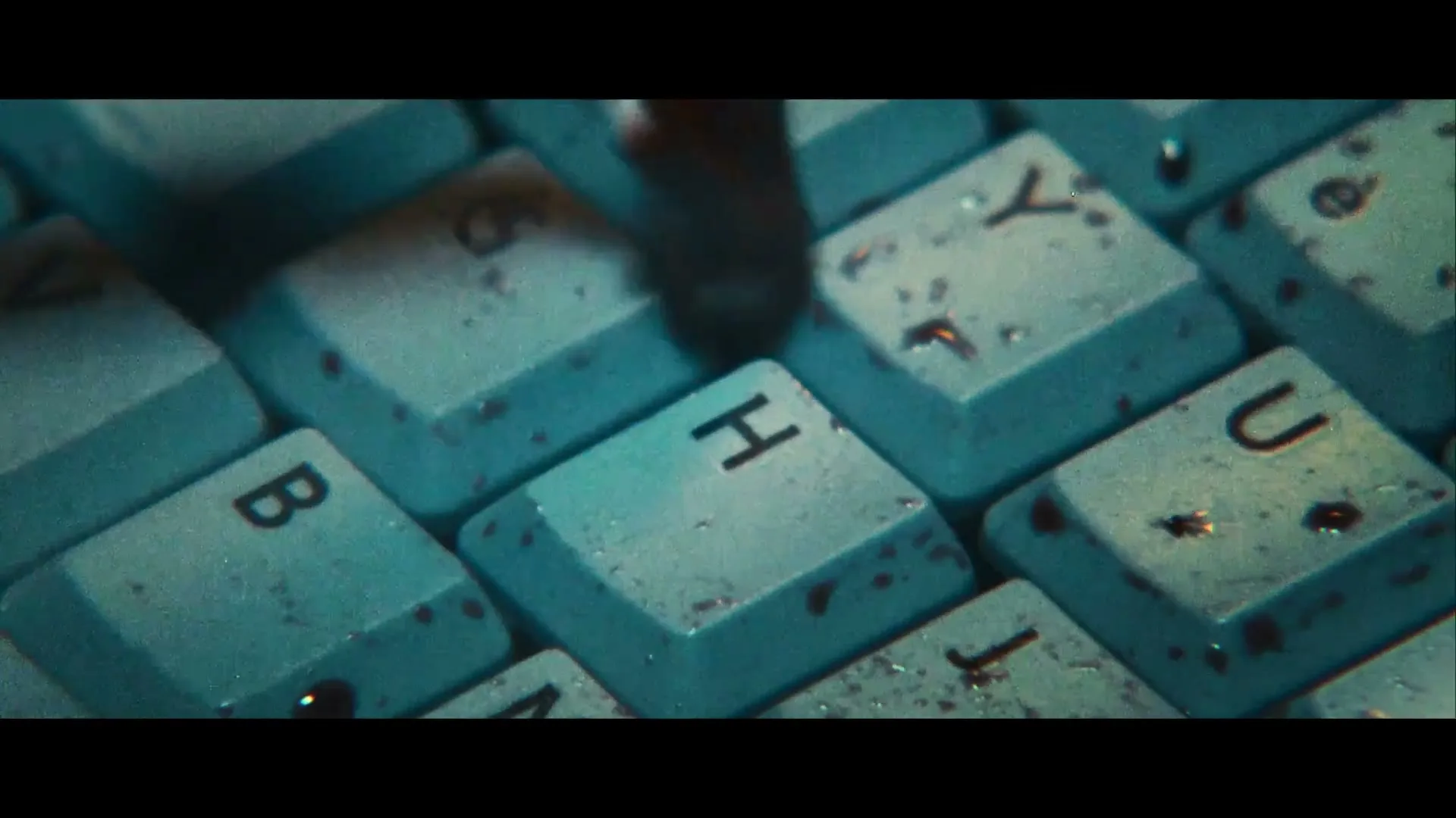 A finger presses the 'H' key on a dirty, teal-colored computer keyboard, with other keys visible around it.