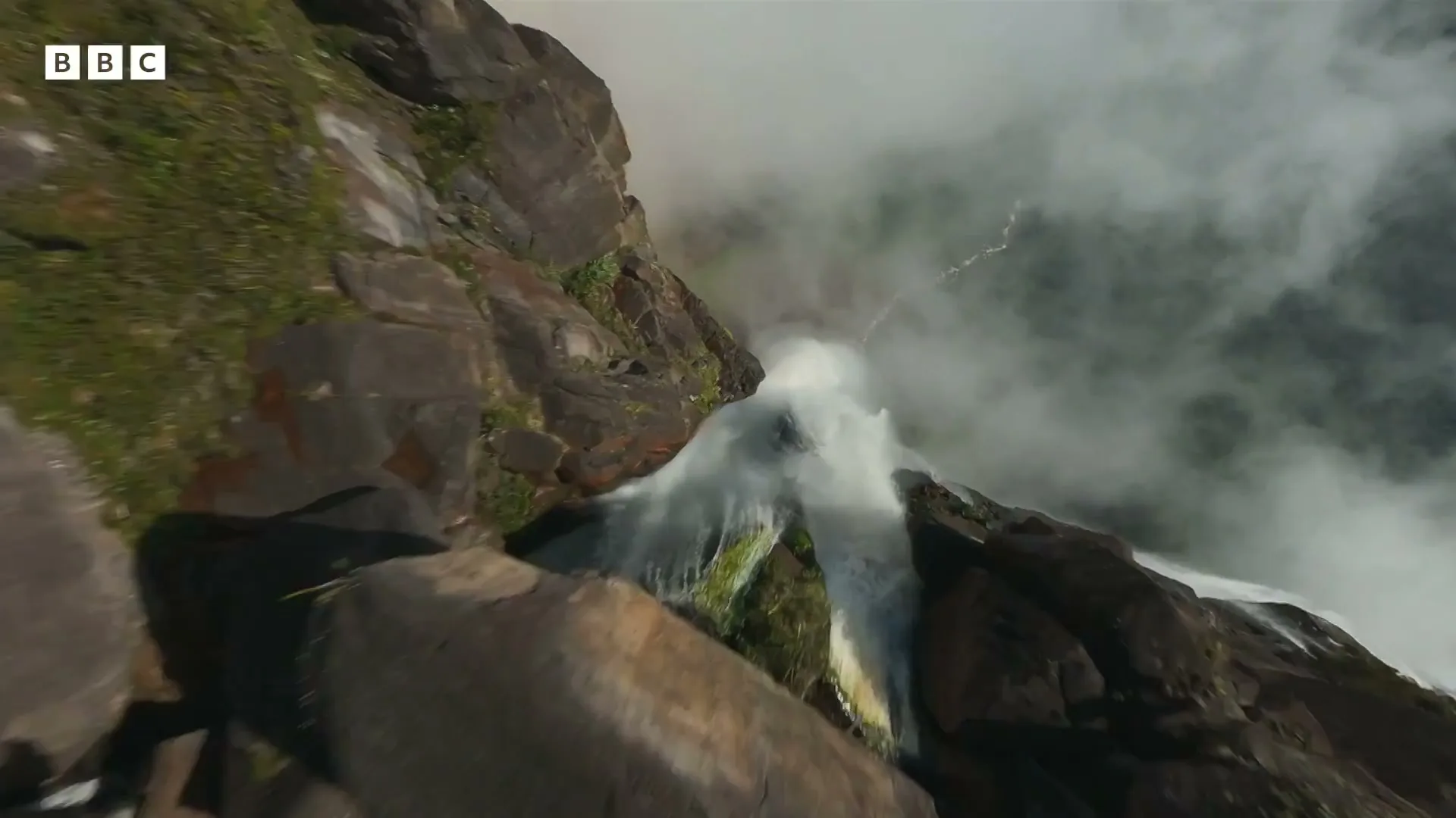 An extreme wide shot from above shows a massive waterfall cascading down a rocky cliff face, surrounded by mist and clouds.