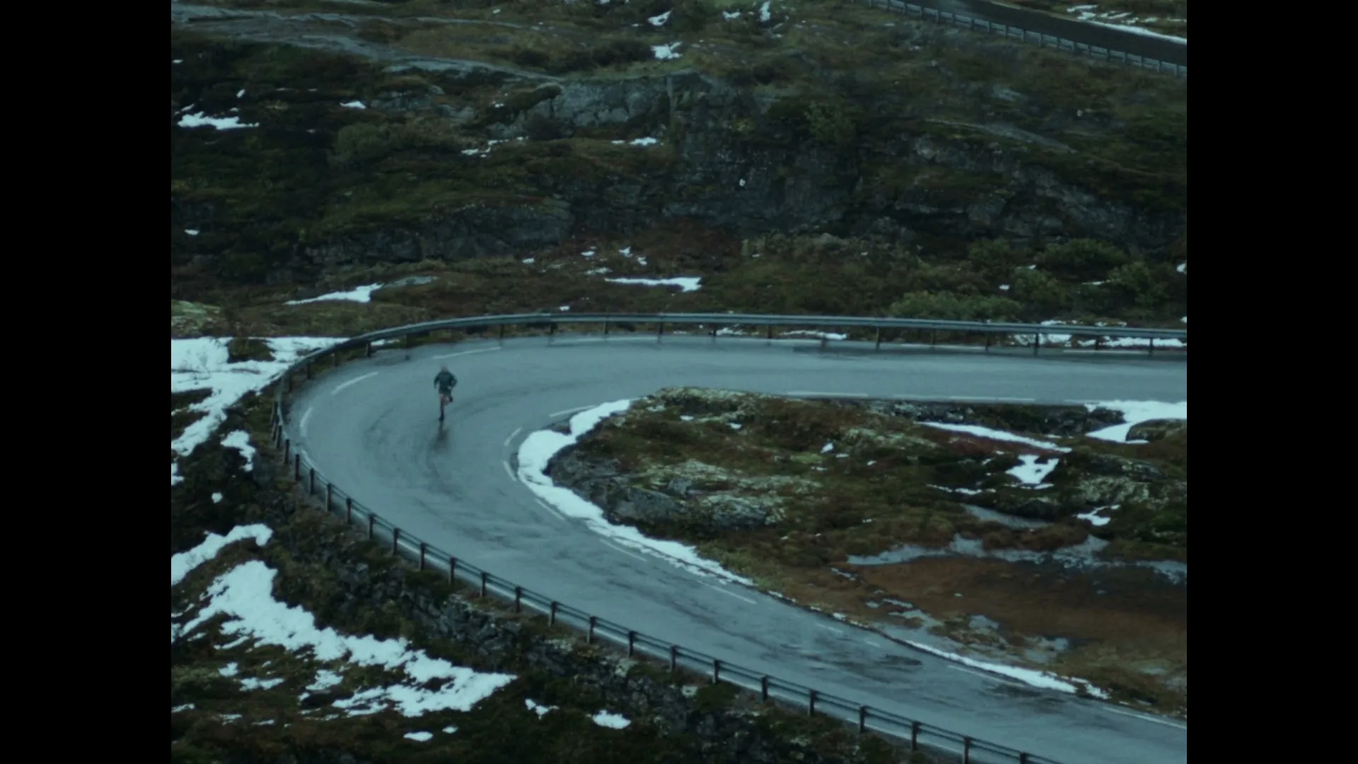 A lone male cyclist in athletic wear rides a winding, wet mountain road with patches of snow and green moss, under an overcast sky.