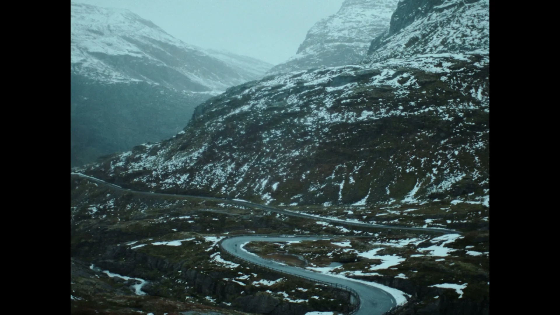 A winding road snakes through a snow-dusted mountain pass under a grey sky, with no people or vehicles visible.