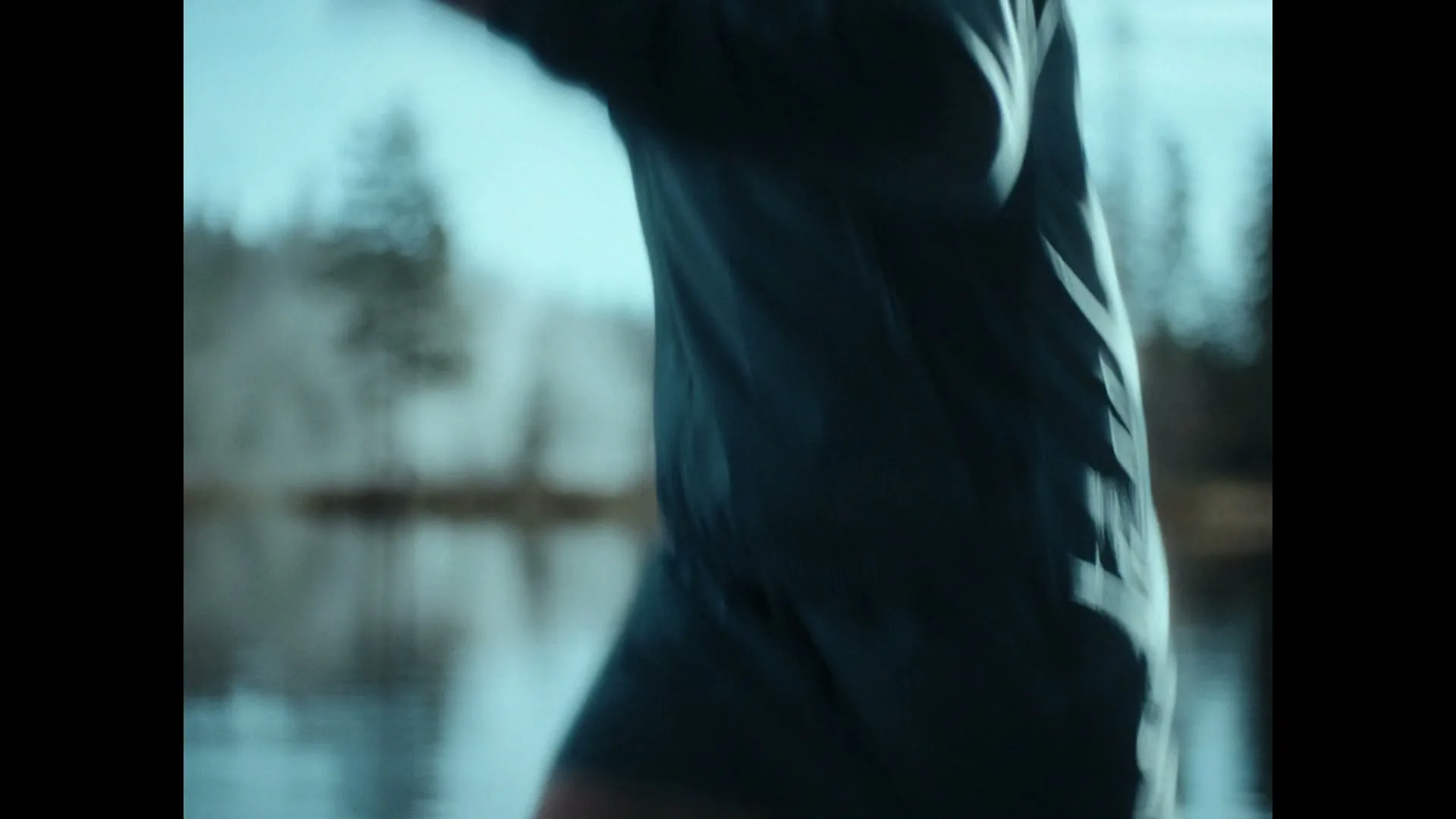 A male athlete with a man bun and earbuds runs across a log over a body of water in a forest.