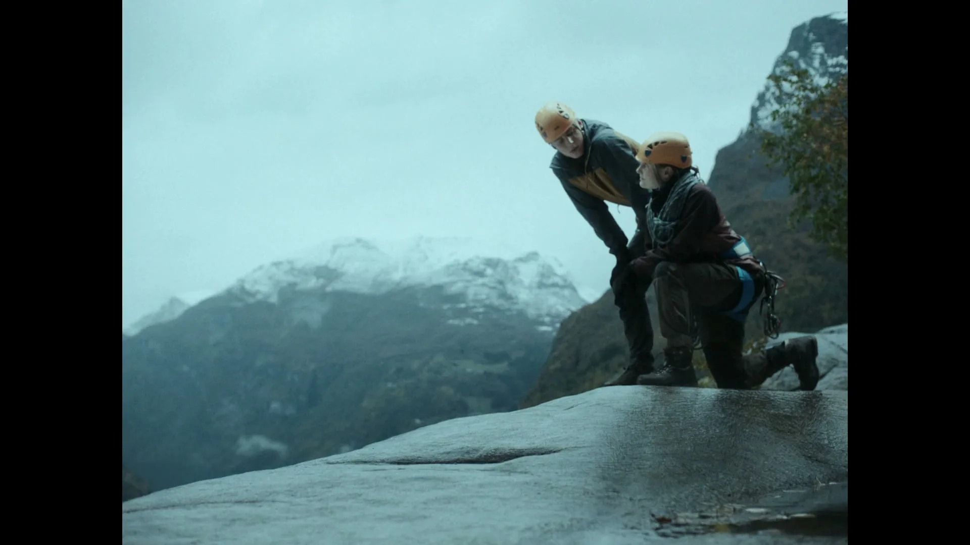 Two people in climbing gear and helmets look down from a cliff edge at a snowy mountain range in the distance.