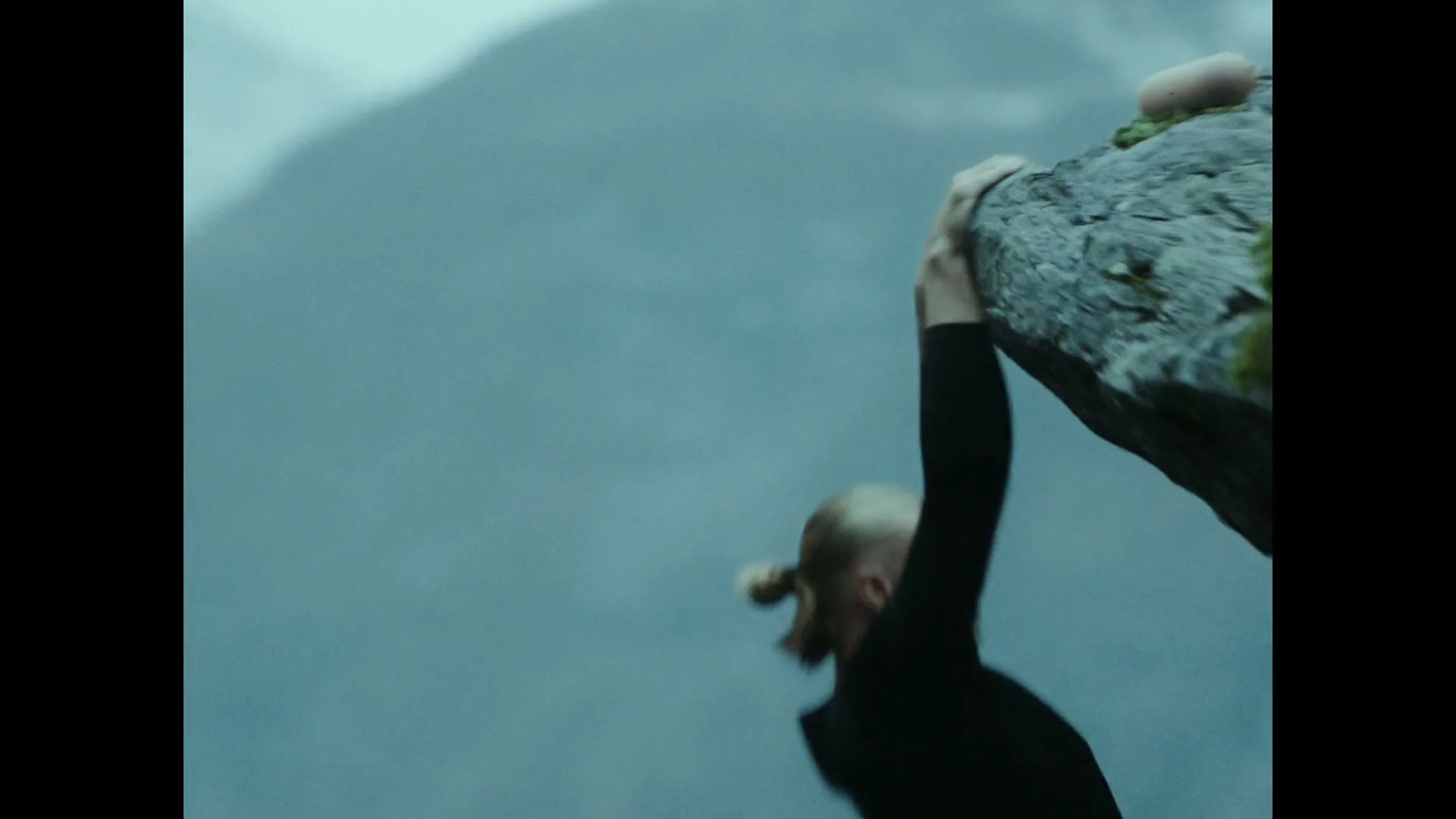 A male rock climber with a blonde top knot hangs precariously from a cliff edge, overlooking a vast fjord and snow-capped mountains on a cloudy day.