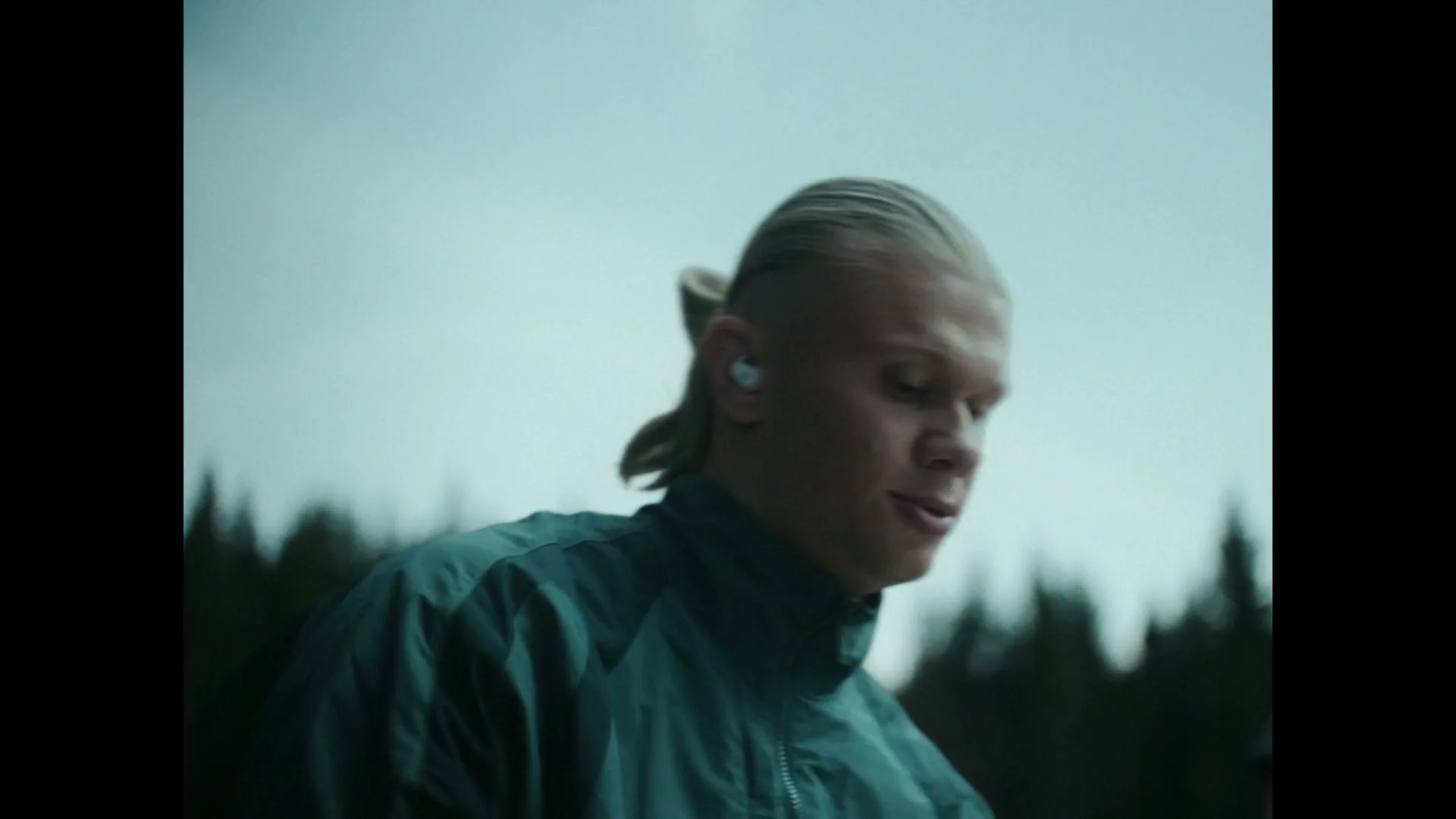 A young man with a blonde ponytail and a teal jacket swings an axe, wearing white earbuds, against a cloudy sky and forest background.