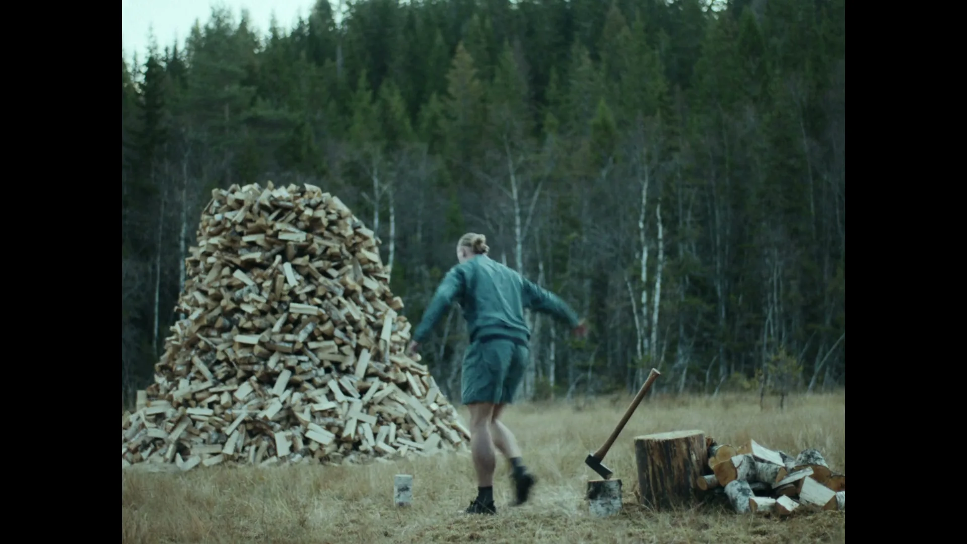 A man in athletic wear throws a piece of wood onto a large pile of firewood in a clearing surrounded by a forest.