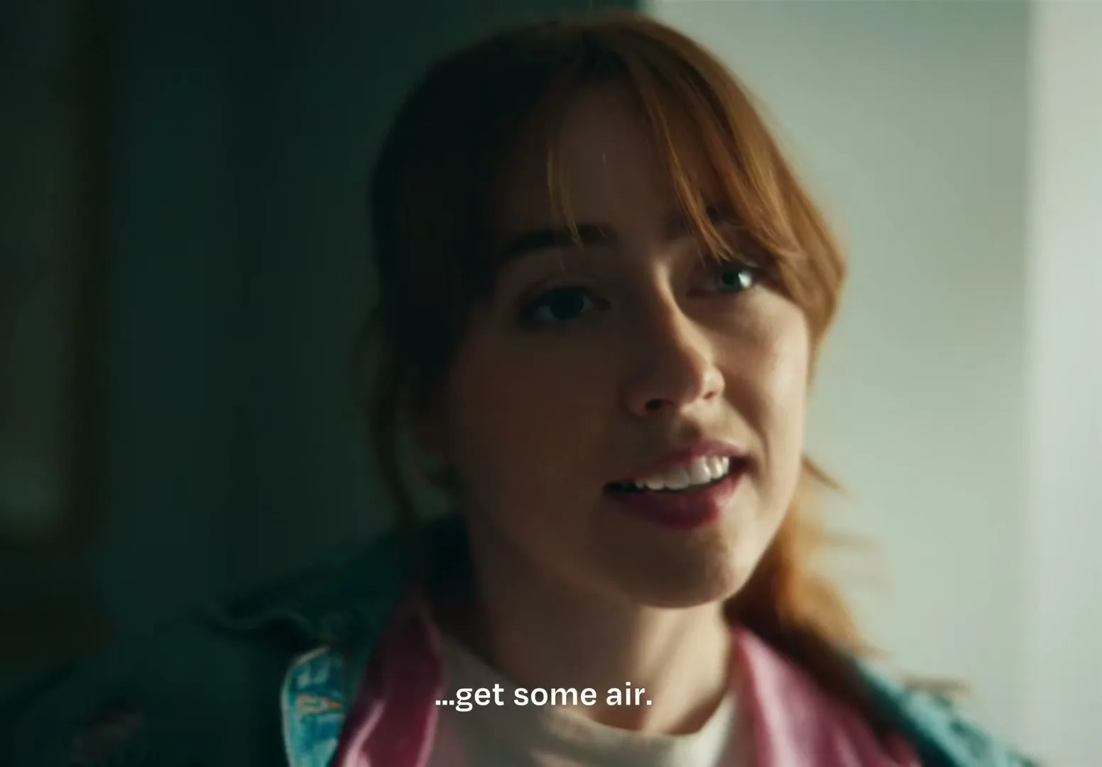 A young woman with red hair, wearing a pink shirt and denim jacket, speaks to someone off-camera in a dimly lit room.