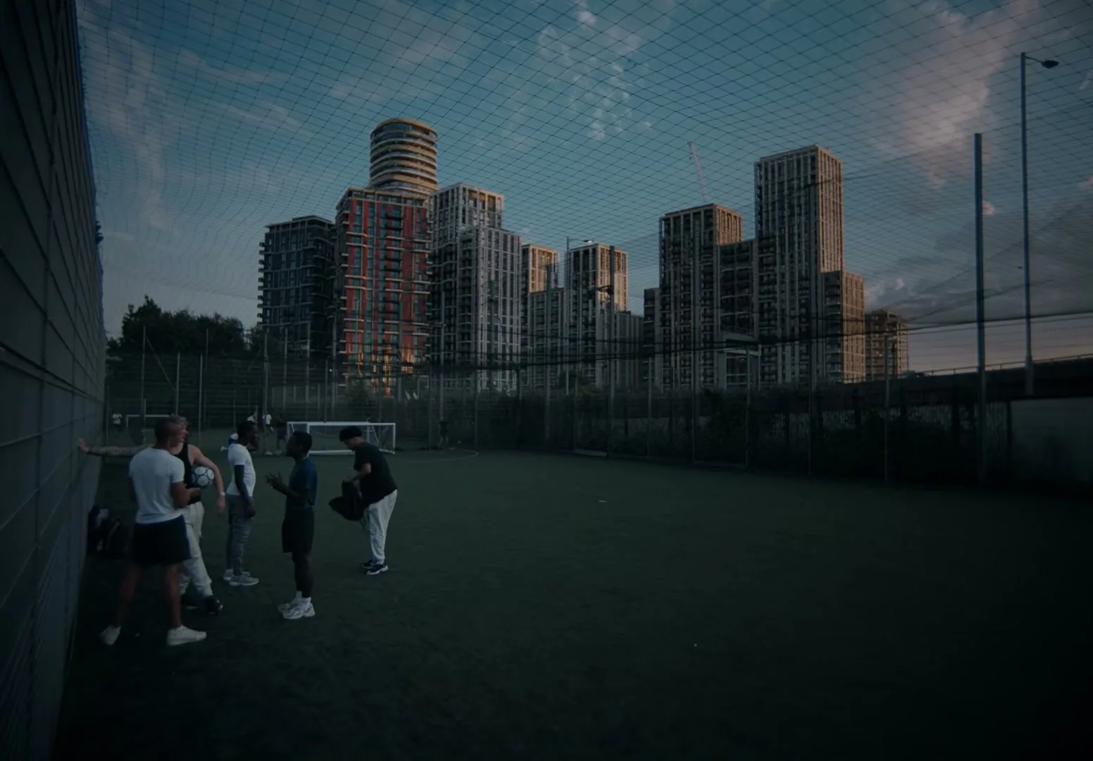 A group of young men are gathered on a football pitch at dusk, with tall residential buildings in the background, as one of them prepares to leave.
