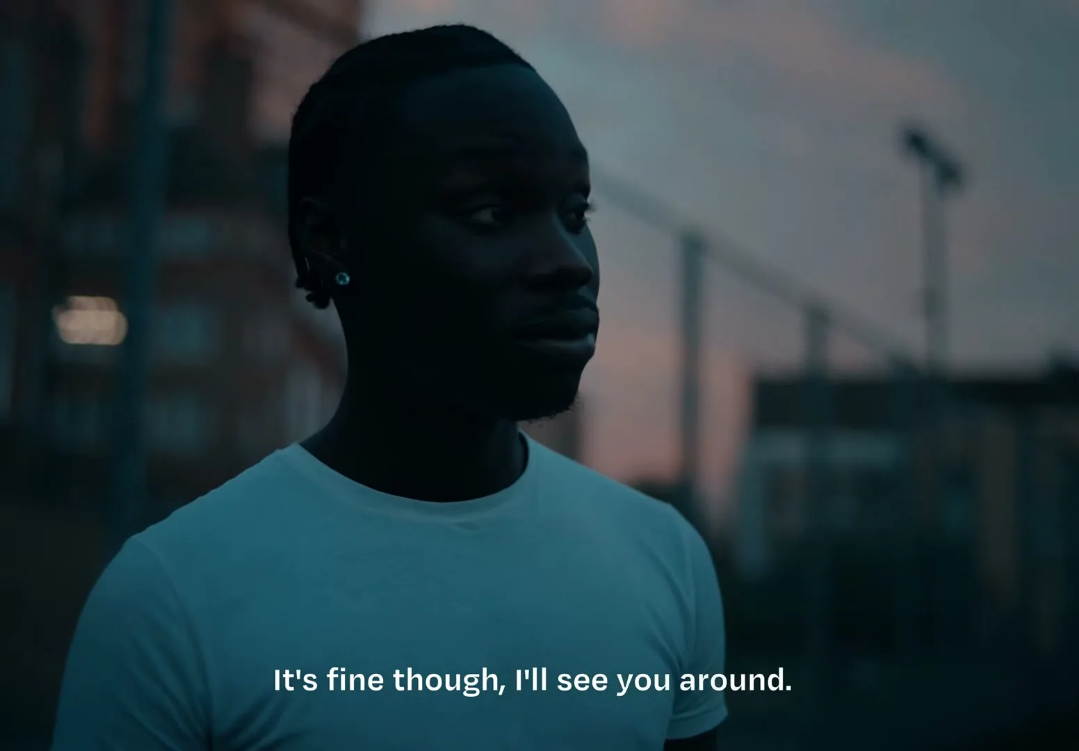 A young Black man in a white t-shirt looks around with a neutral expression in an urban outdoor setting at dusk.