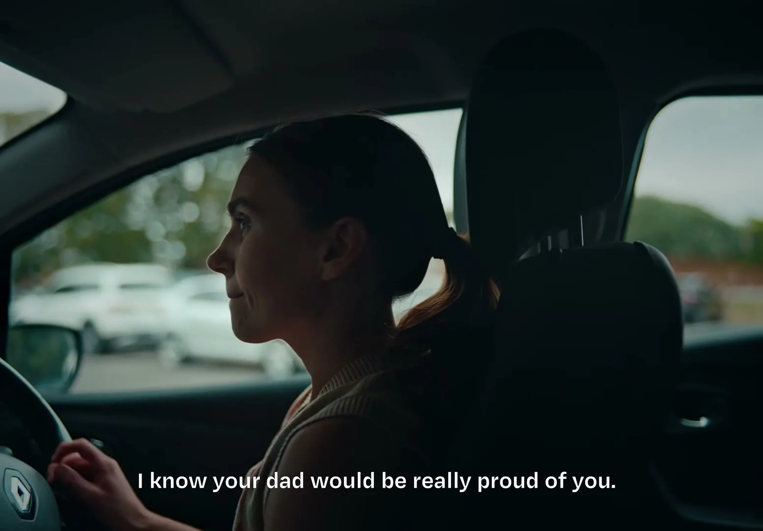 A young woman with dark hair in a ponytail sits in the driver's seat of a car, looking out the window with a neutral expression.