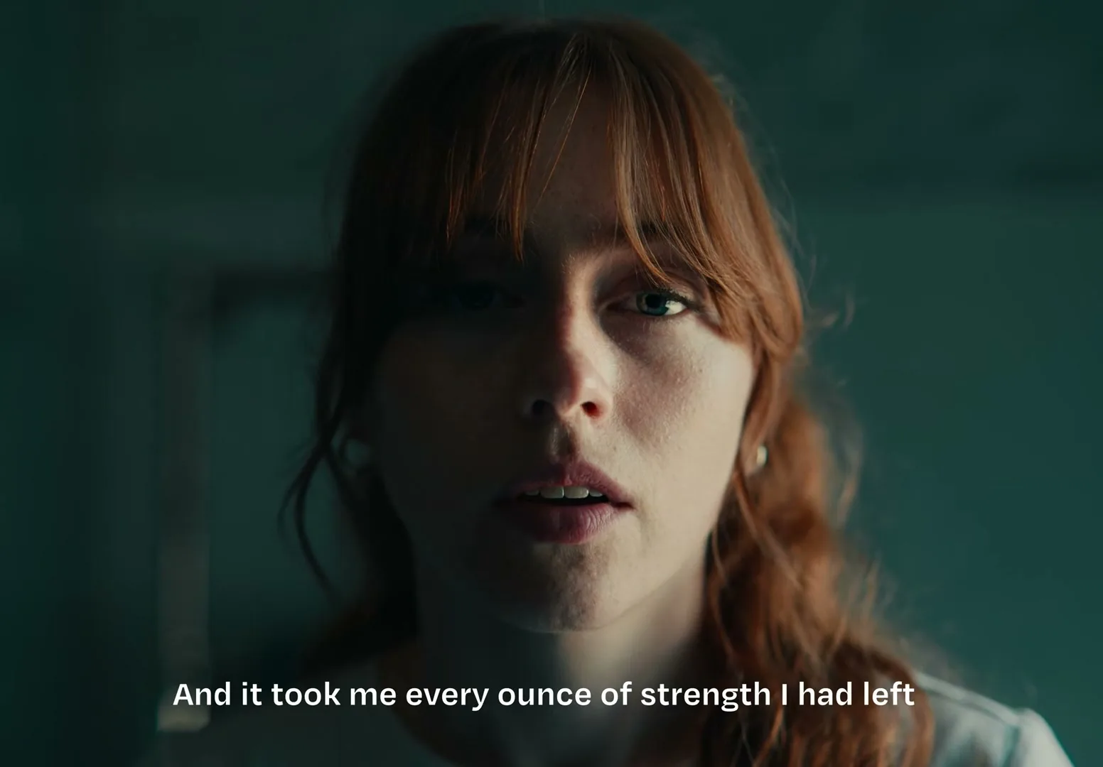A young woman with red hair and a white t-shirt looks directly at the camera, her face partially shadowed, in an interior setting.