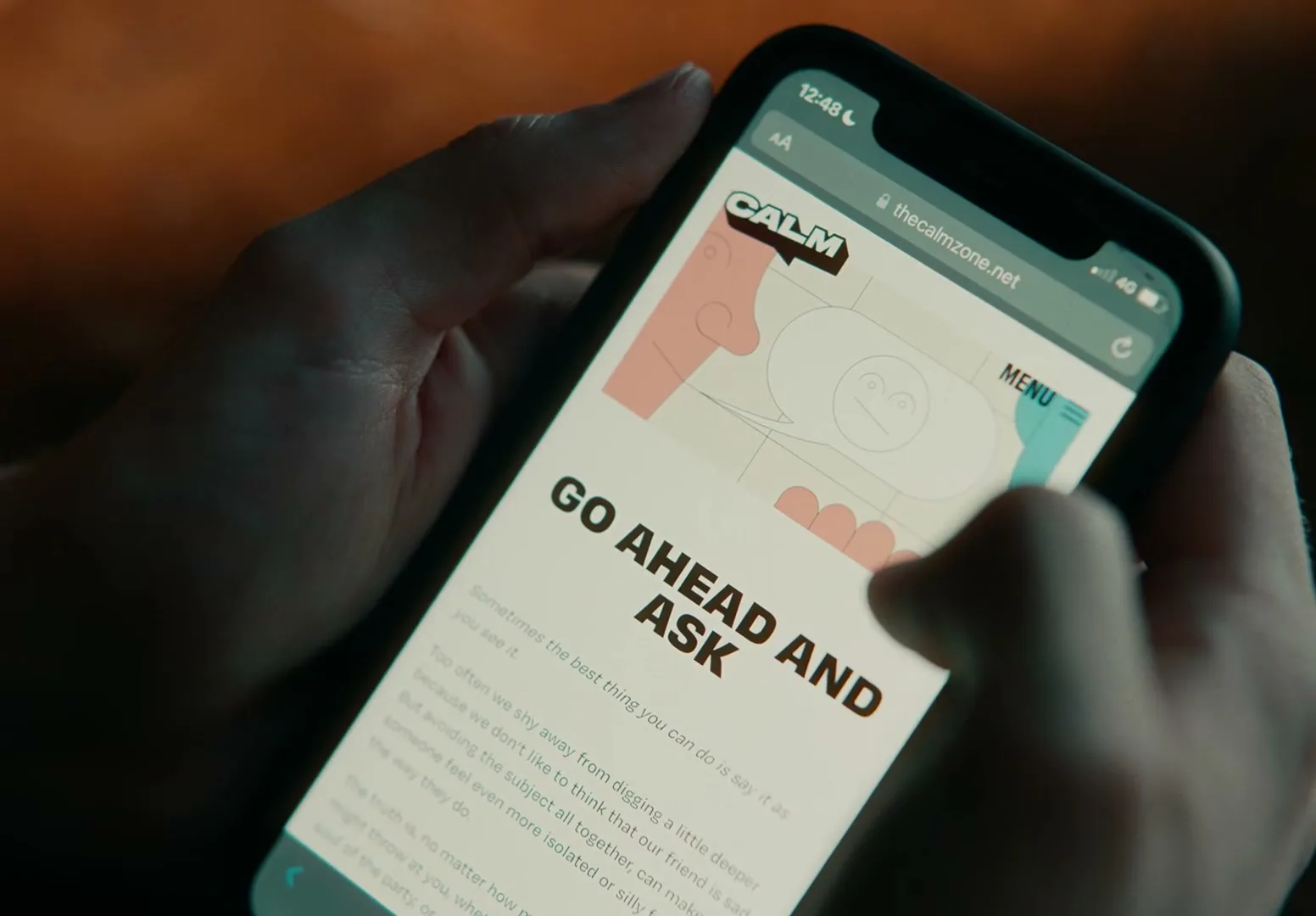 A person holds a smartphone displaying the CALM website with the text "GO AHEAD AND ASK" on screen, in an interior setting.