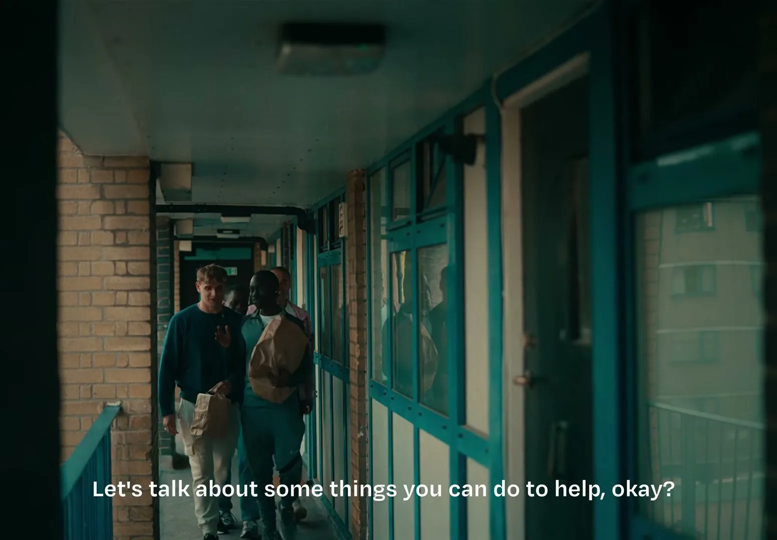 A group of young men walk down a narrow, brick-walled apartment building hallway, carrying paper bags, as they talk to each other.
