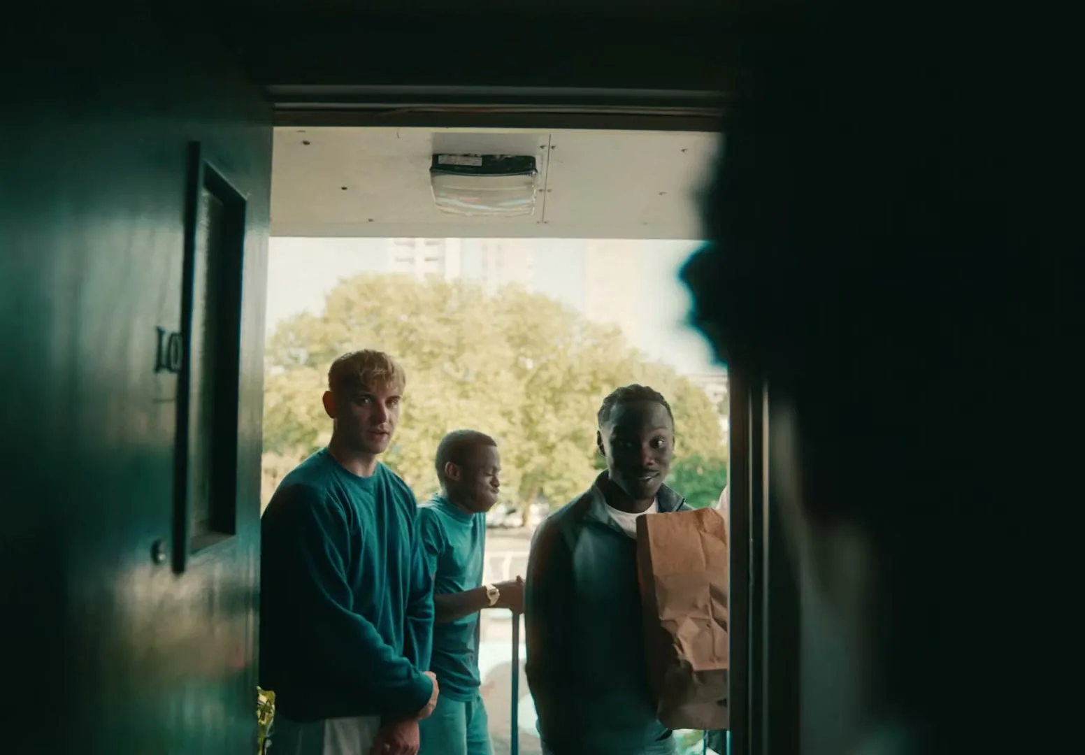 Three young men stand at an open doorway, looking into a dark room, with one holding a paper bag, in a residential building.