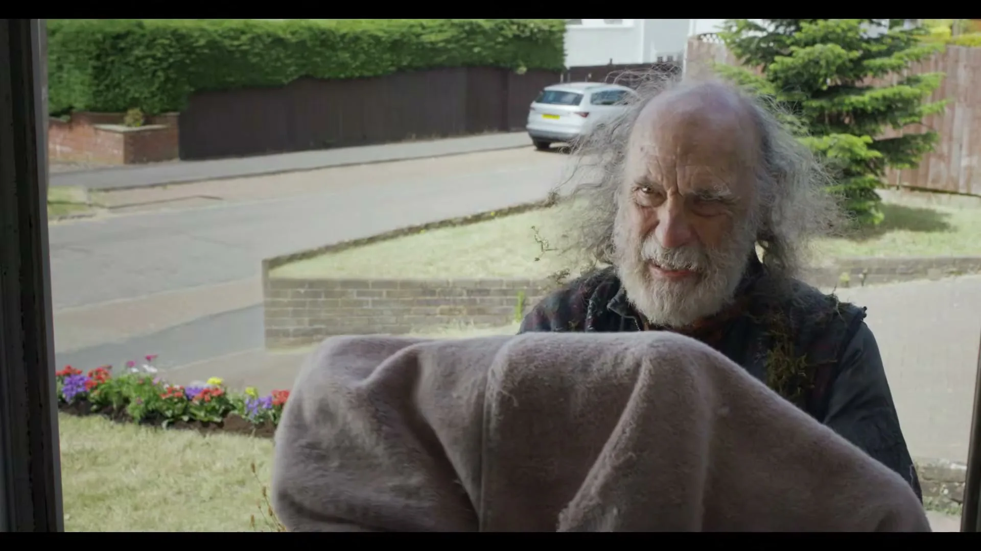 A disheveled older man with a white beard and wild hair stands outside a house, holding a blanket and winking, as seen through a window.