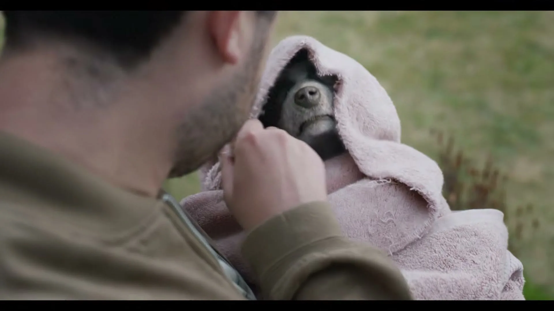 A man holds a small, dark-furred animal wrapped in a pink towel, gently stroking its head outdoors in daylight.