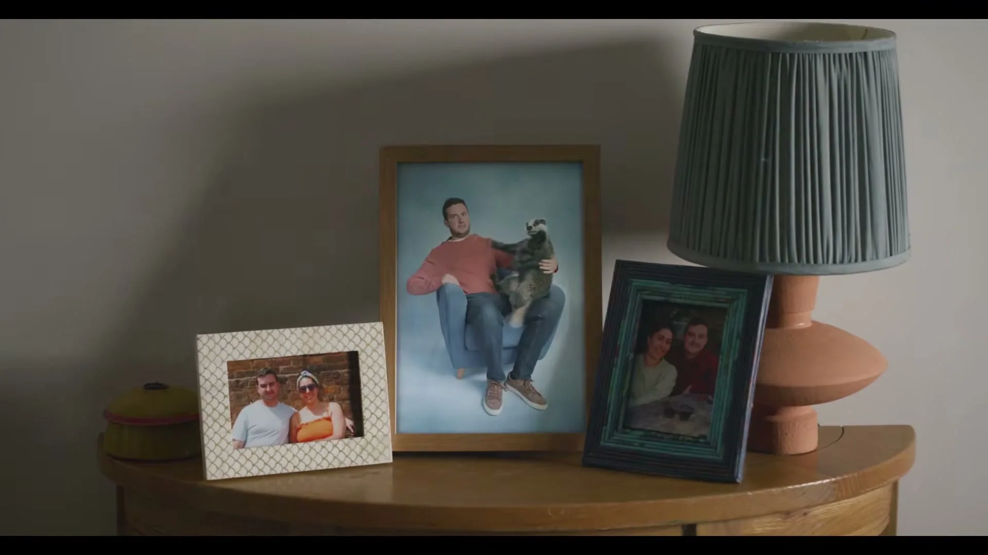 Three framed photos on a wooden table, with a lamp on the right, show a man with a badger, a couple, and another couple, as the man in the middle photo changes clothes.
