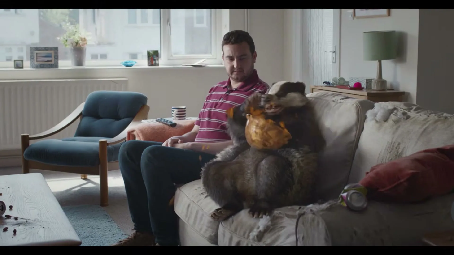 A man sits on a sofa next to a badger eating crisps from a bowl, making a mess in a messy living room.