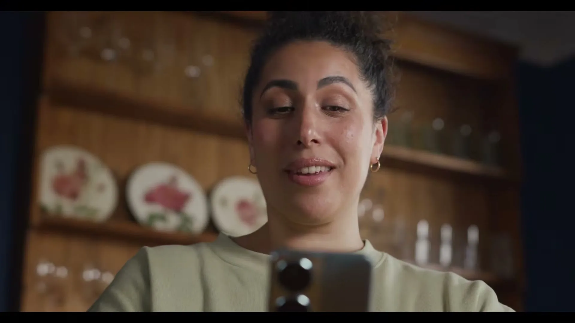 A young woman with curly hair in a light green top looks at her phone and smiles, in a kitchen with wooden shelves.