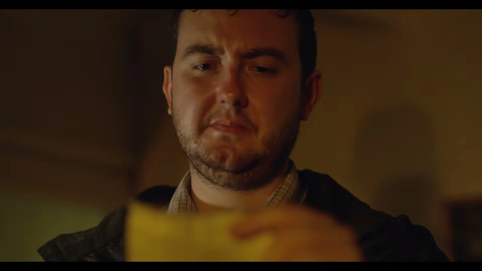A man with a beard looks down at a yellow paper, his face showing a mix of sadness and concern in a dimly lit interior.