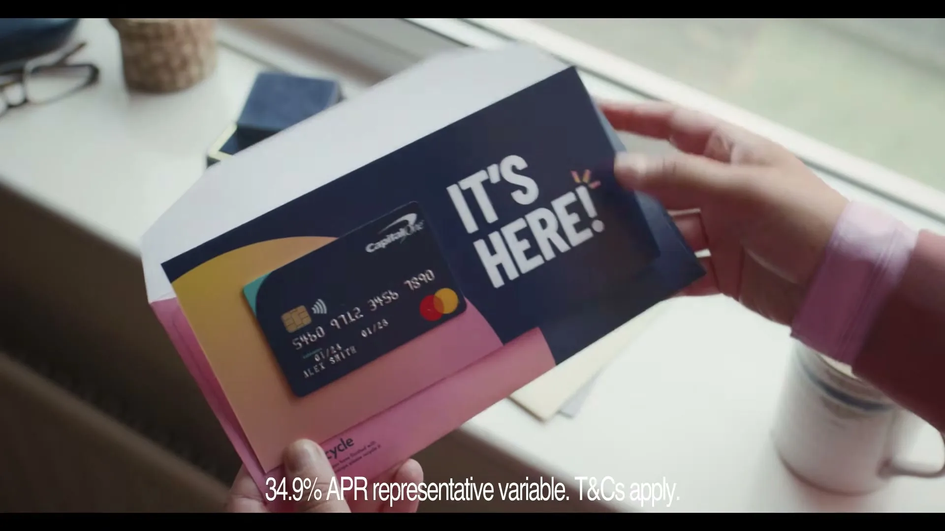 A person's hands hold a pink envelope, revealing a Capital One credit card and a card with "IT'S HERE!" written on it, next to a window.