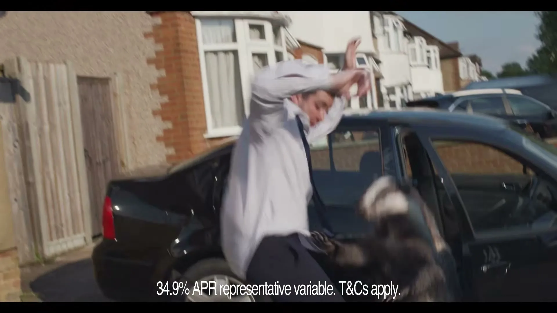 A man in a white shirt and dark trousers is surprised by a badger jumping out of a black car in a suburban street.