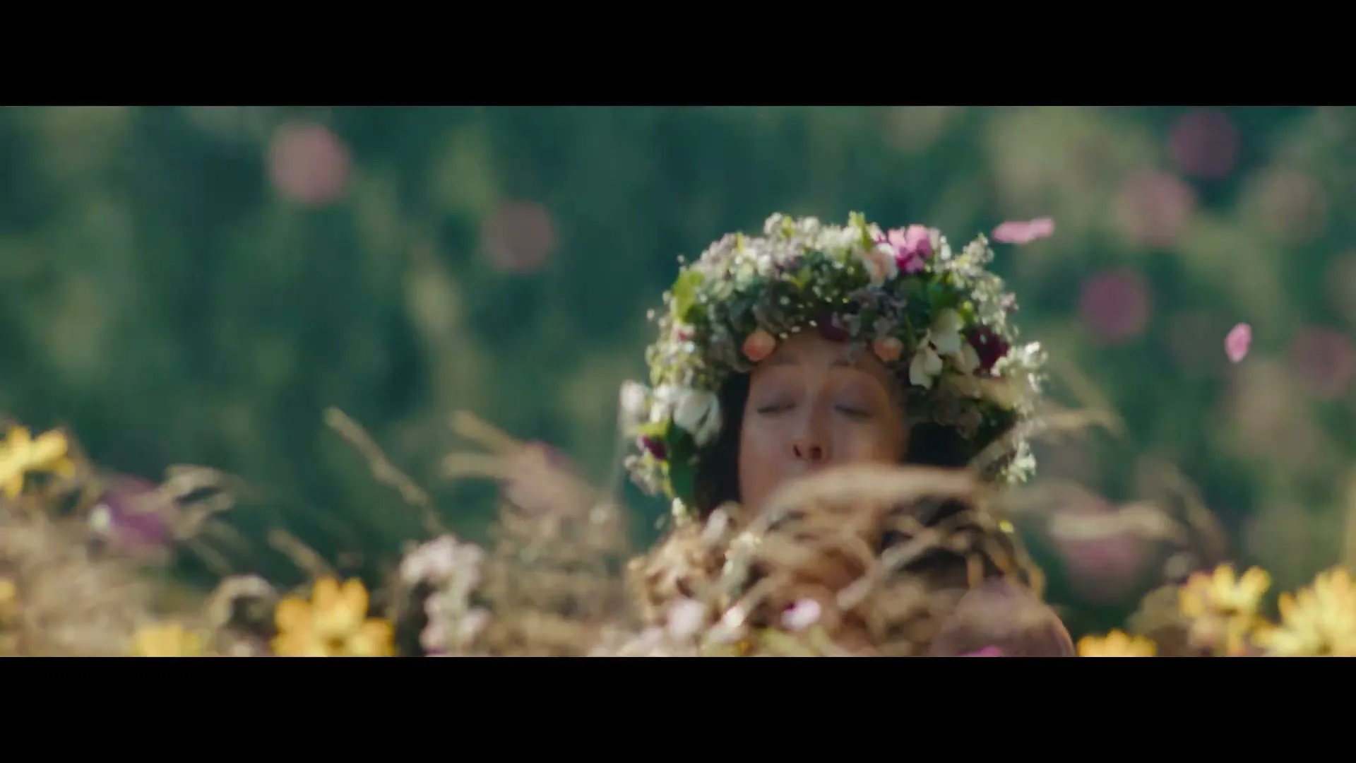 A woman with a flower crown plays a saxophone in a field of wildflowers, then a falcon is seen perched with pink petals falling around it.