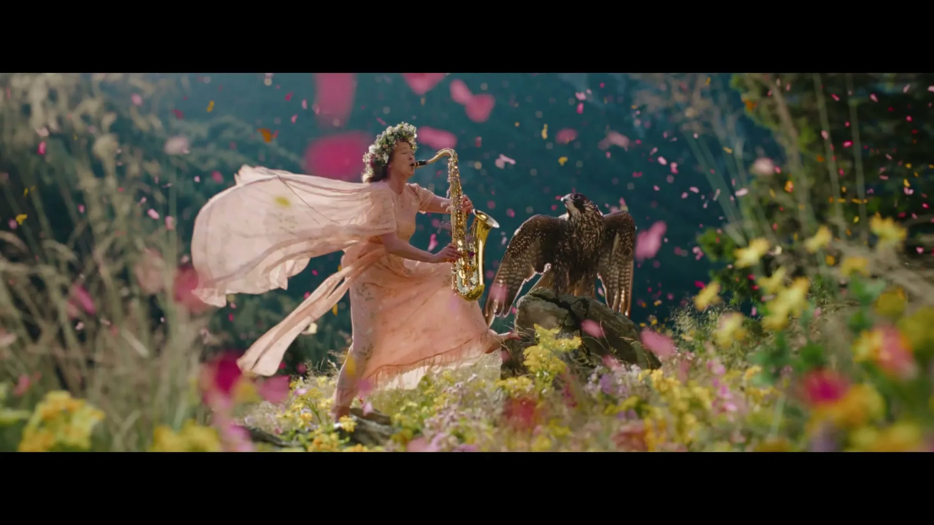 A woman in a flowing pink dress and flower crown plays a saxophone next to a falcon on a rock, surrounded by falling petals and wildflowers.