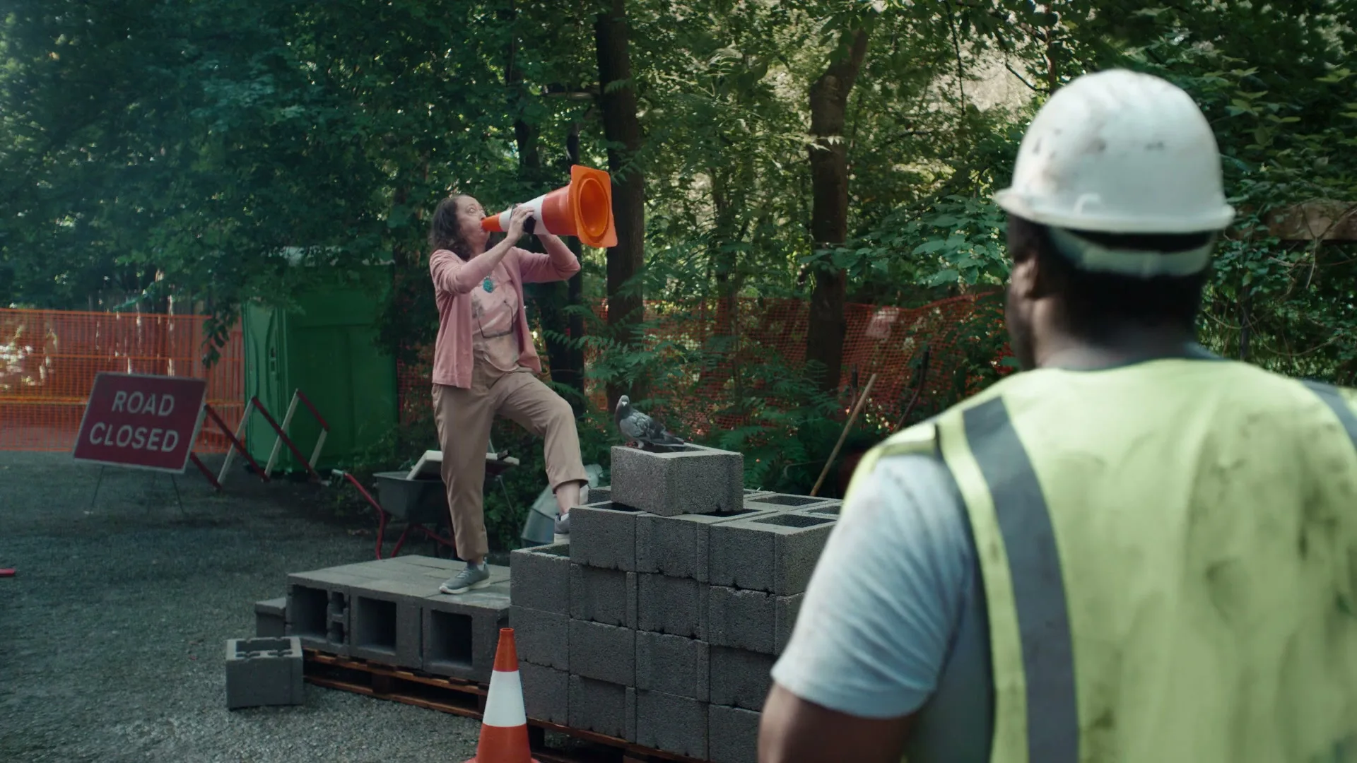 A woman in a pink shirt and beige pants shouts into an orange traffic cone at a construction worker, who is seen from behind, at a construction site.