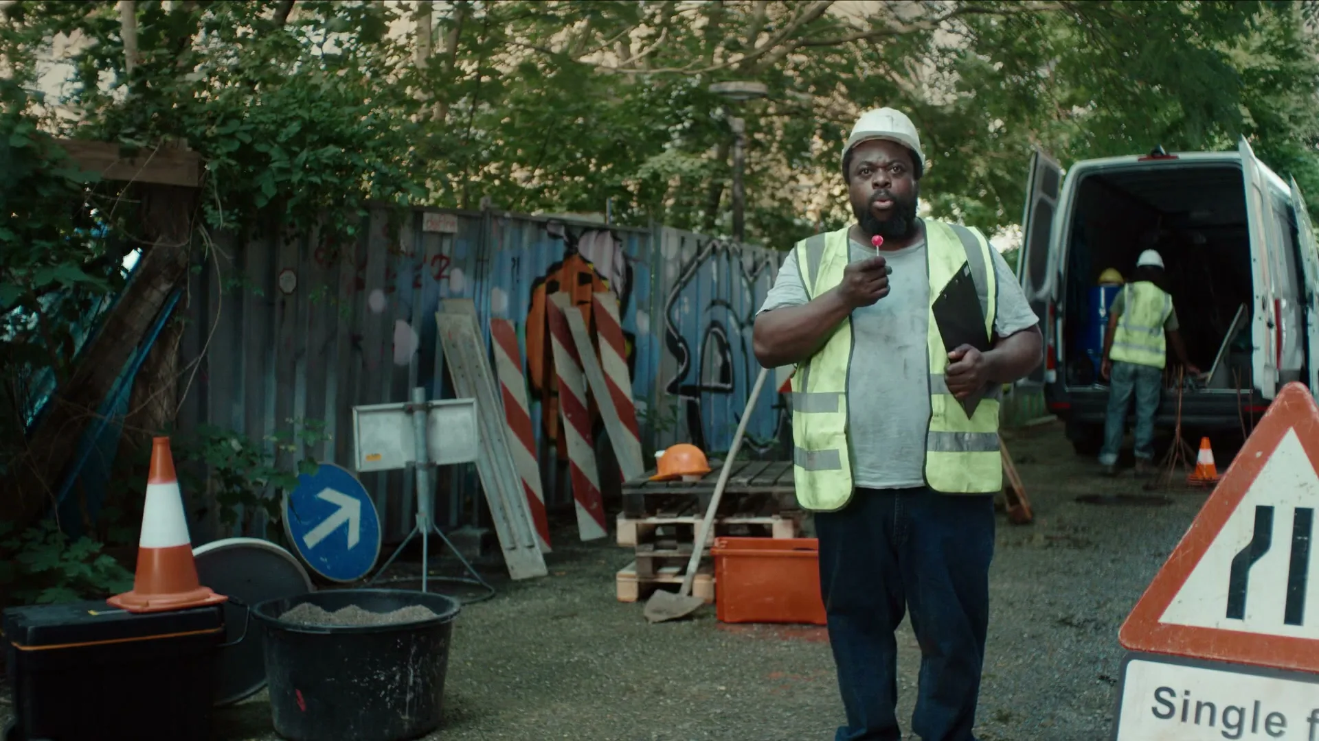 A construction worker in a hard hat and high-vis vest holds a clipboard and a lollipop, looking directly at the camera on a construction site.