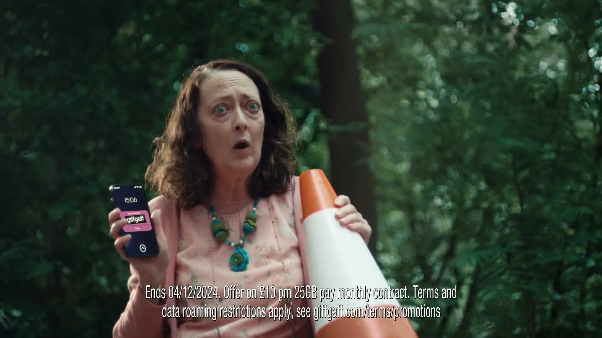 A middle-aged woman with curly brown hair, wearing a pink top and a turquoise necklace, holds a traffic cone and a phone, looking surprised in a park.