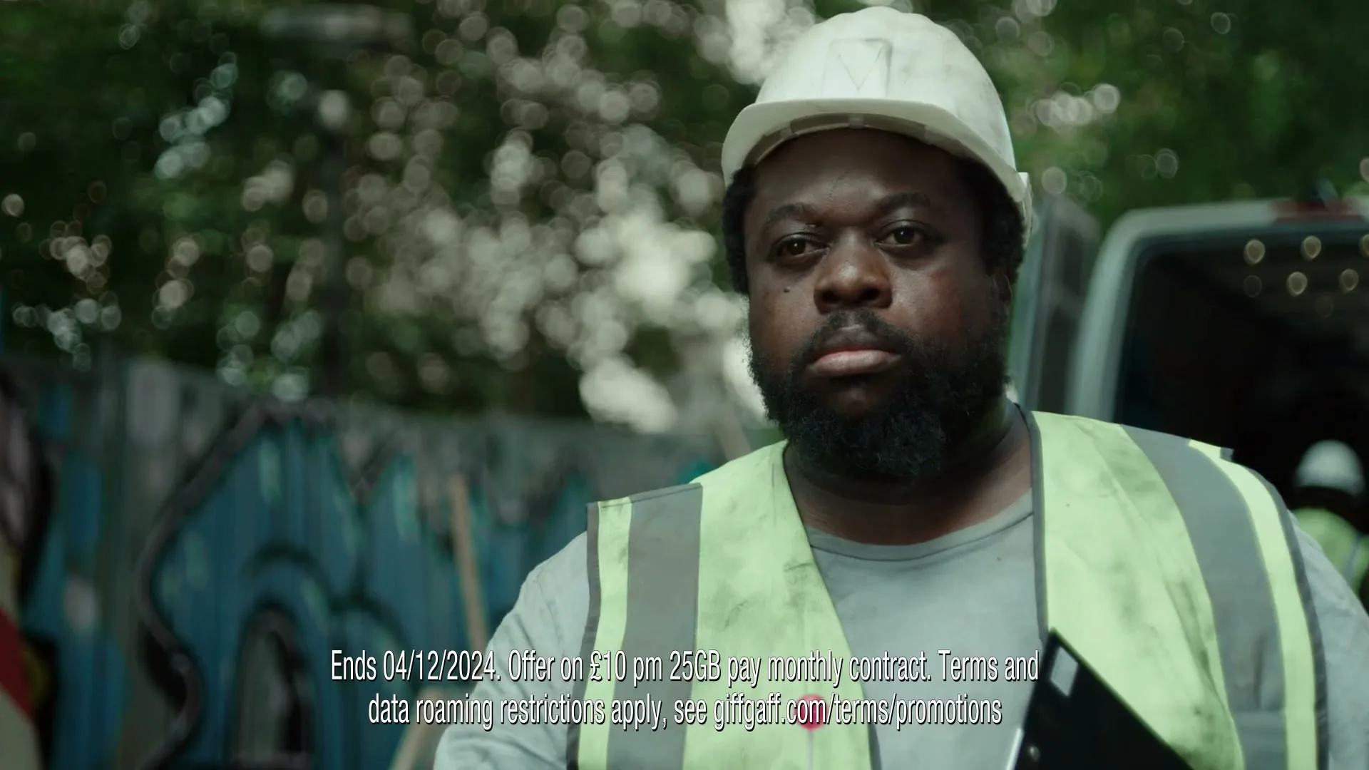 A construction worker in a hard hat and high-vis vest looks directly at the camera with a serious expression in an outdoor setting.